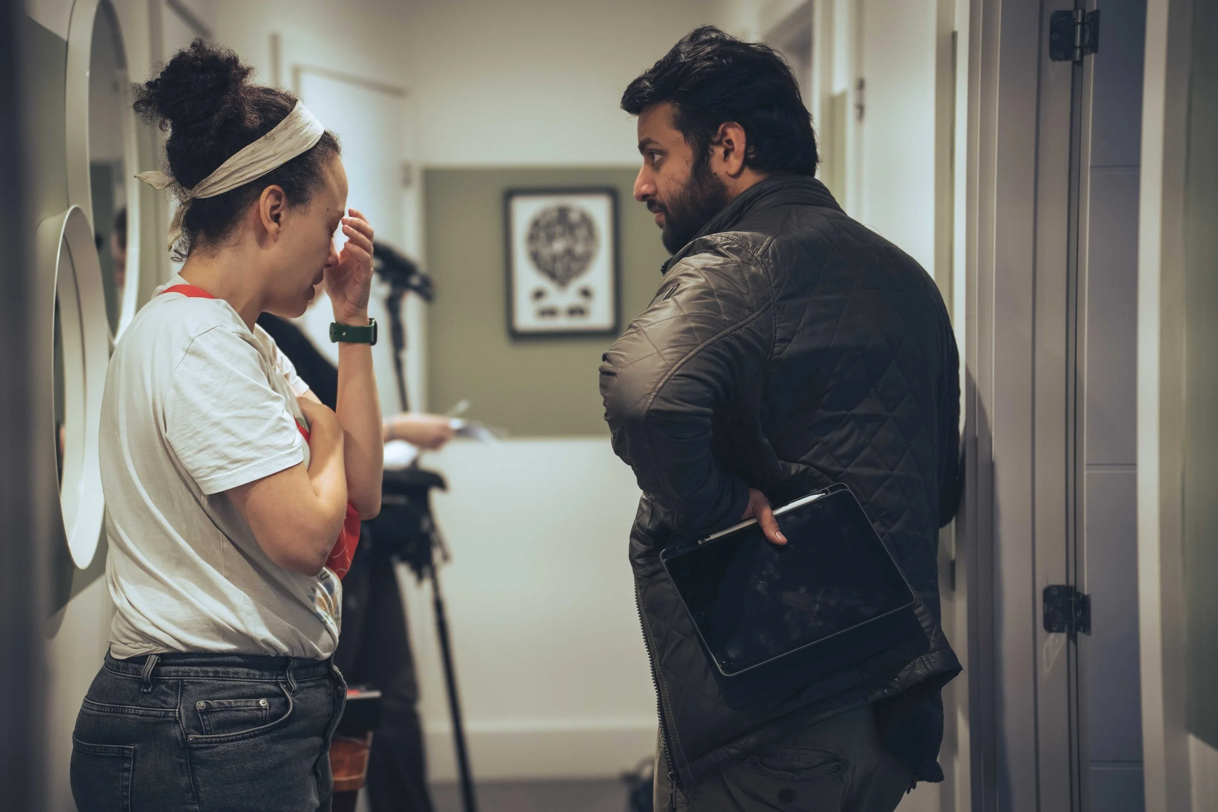 A woman and a man standing in a hallway, engaging in a conversation. The woman appears upset, with her hand on her forehead, while the man is talking to her holdinga folder or a tablet.