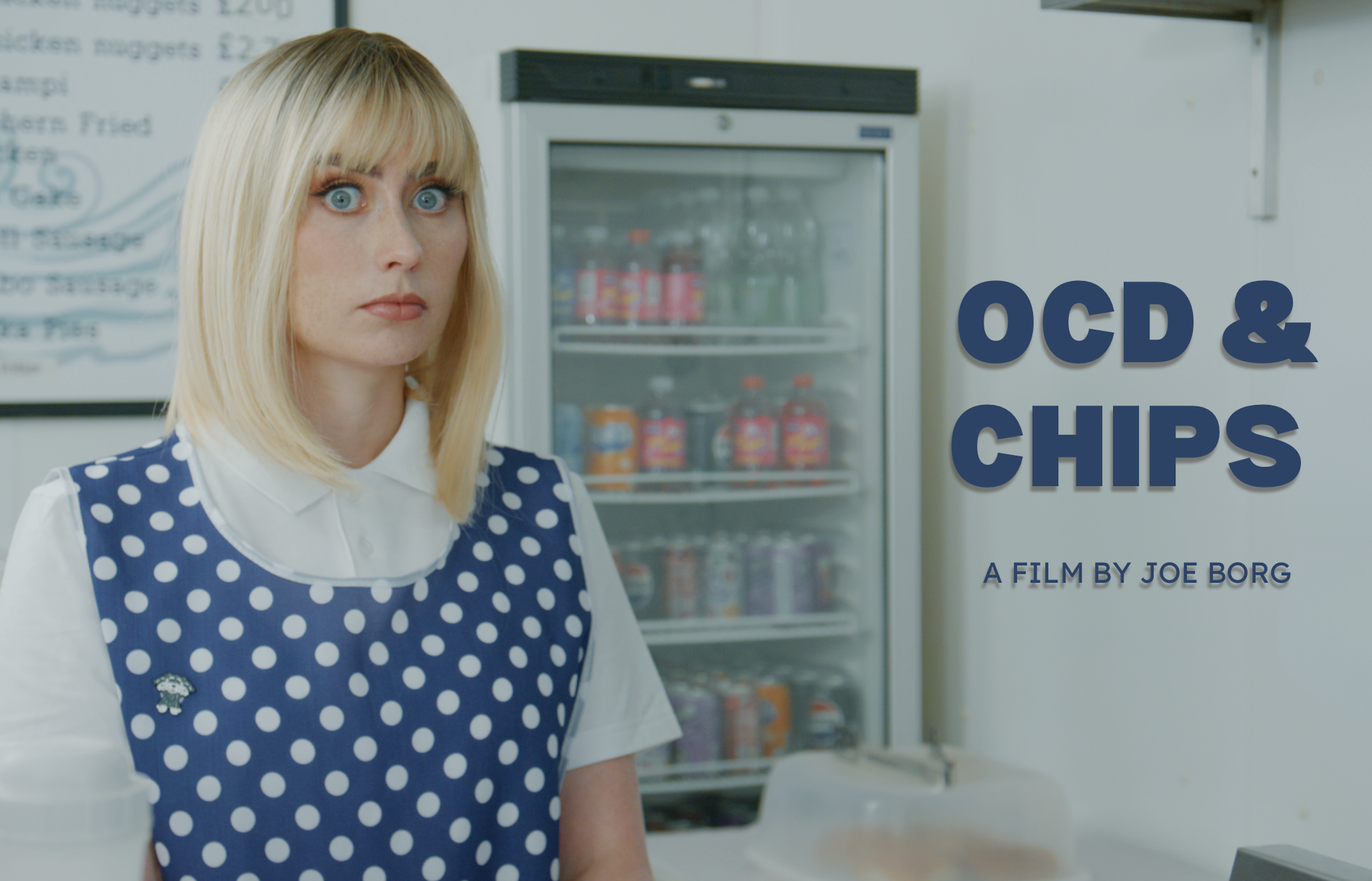 A woman with blonde hair, wearing a blue polka-dot apron over a white shirt, standing in front of a refrigerator in a fast-food restaurant.