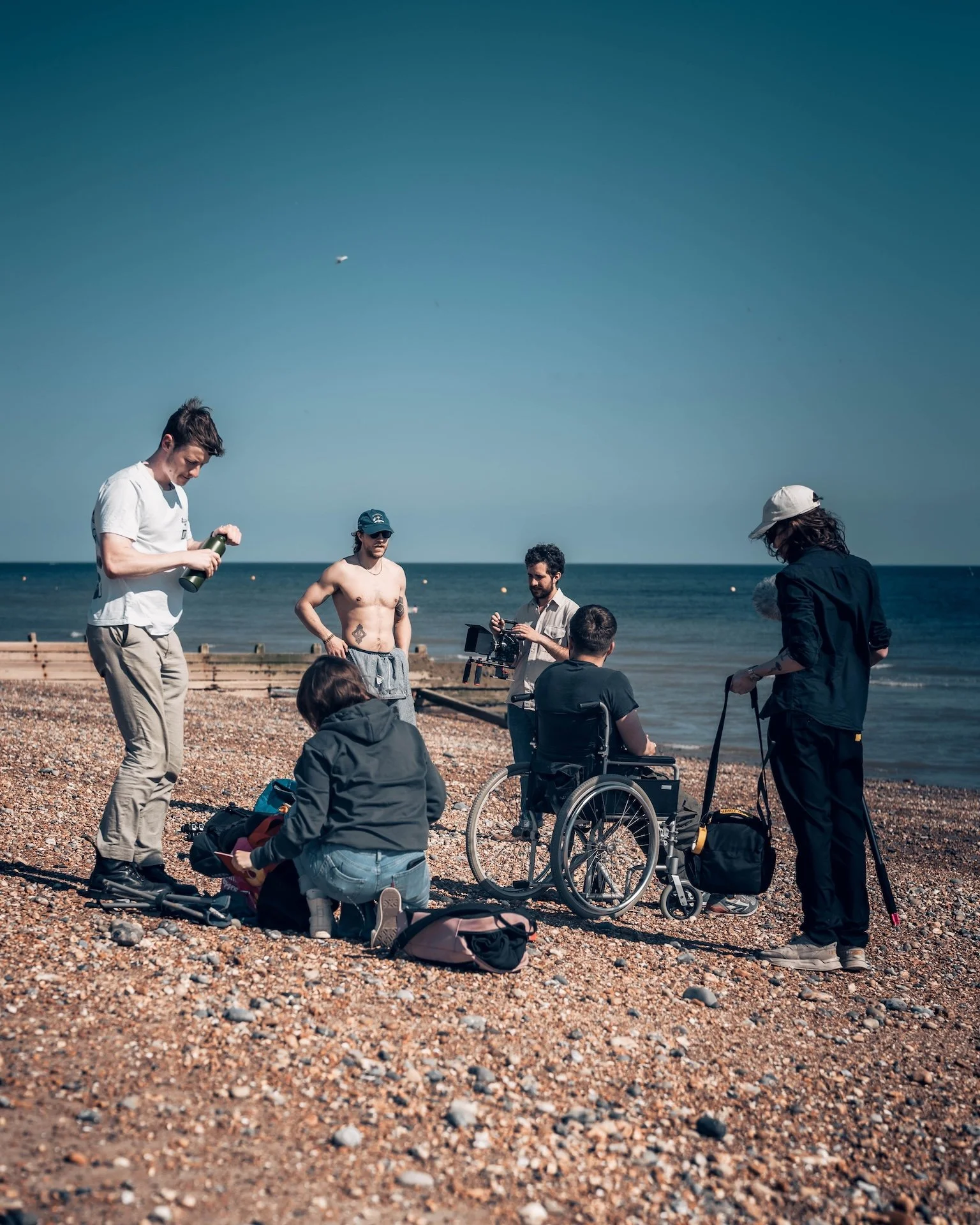 Group of people at the beach, some sitting, some standing, with camera equipment and backpacks, near the shoreline under a clear blue sky.
