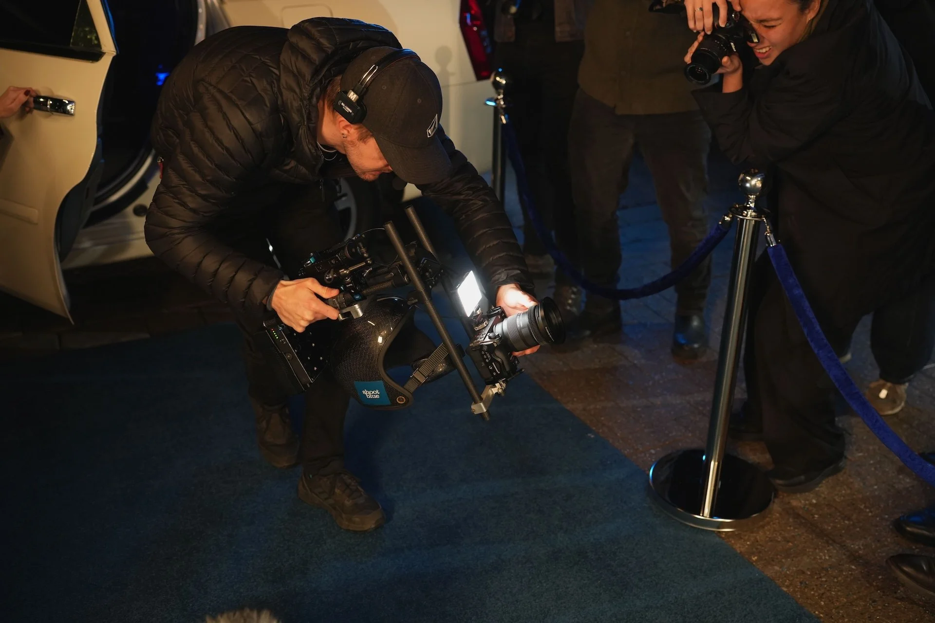 In this photo, a man is crouching while holding a professional camera mounted on a stabilizer rig, aiming it upward. He is wearing a black cap, black headphones, black jacket, and black pants. To the right, a woman is taking a photo with a smaller ca