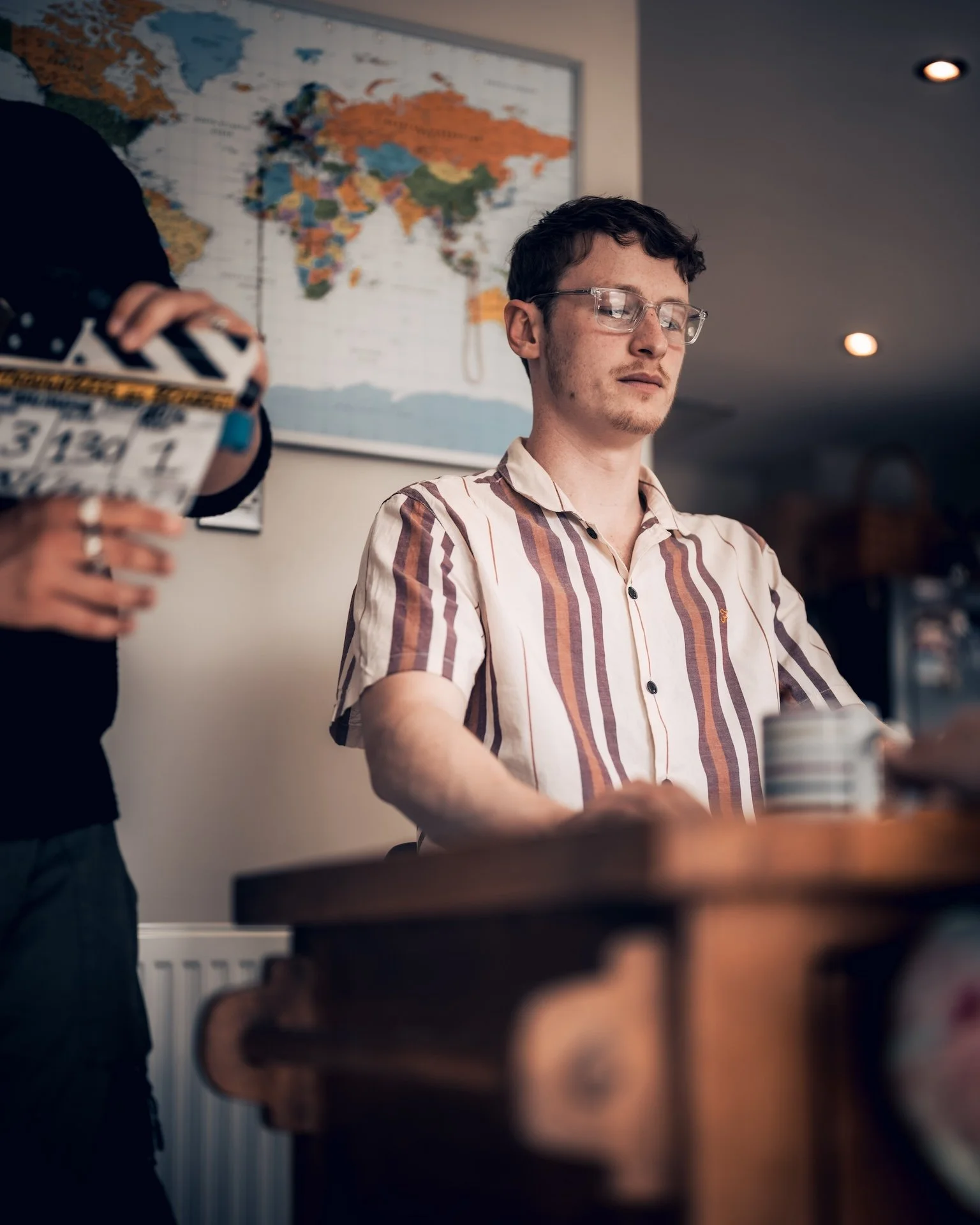 A young man with glasses, wearing a striped shirt, sitting at a table with a drink, in front of a world map, with a person holding a film slate partially visible on the left.