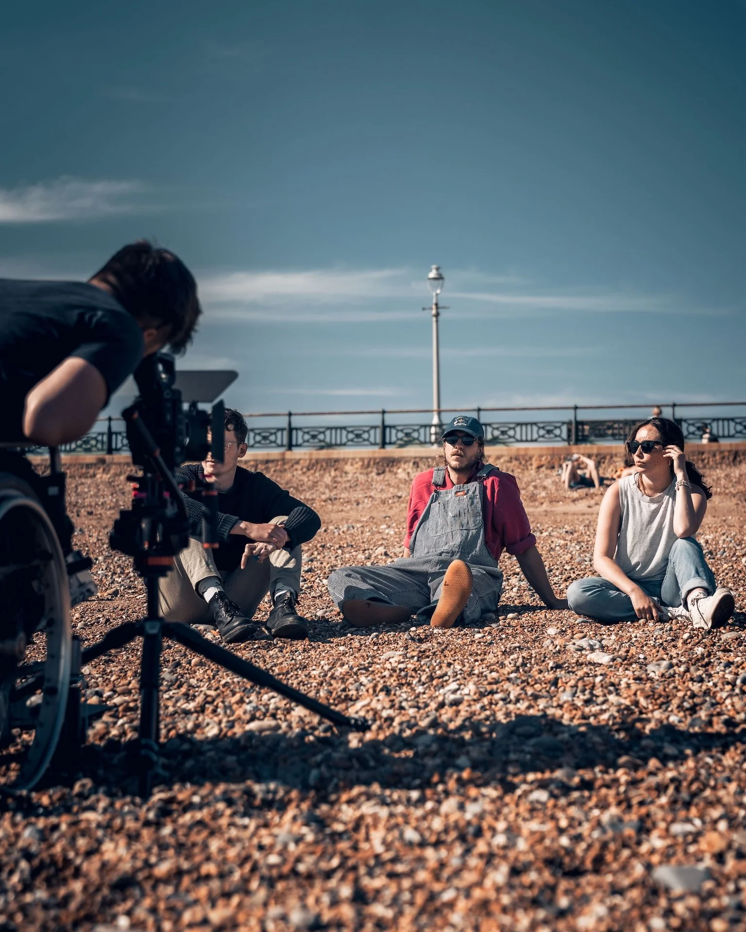 A group of people sitting on a rocky ground near a railing with a lamppost in the background. One person is operating a camera on a tripod, while others are sitting and relaxing, some wearing sunglasses.
