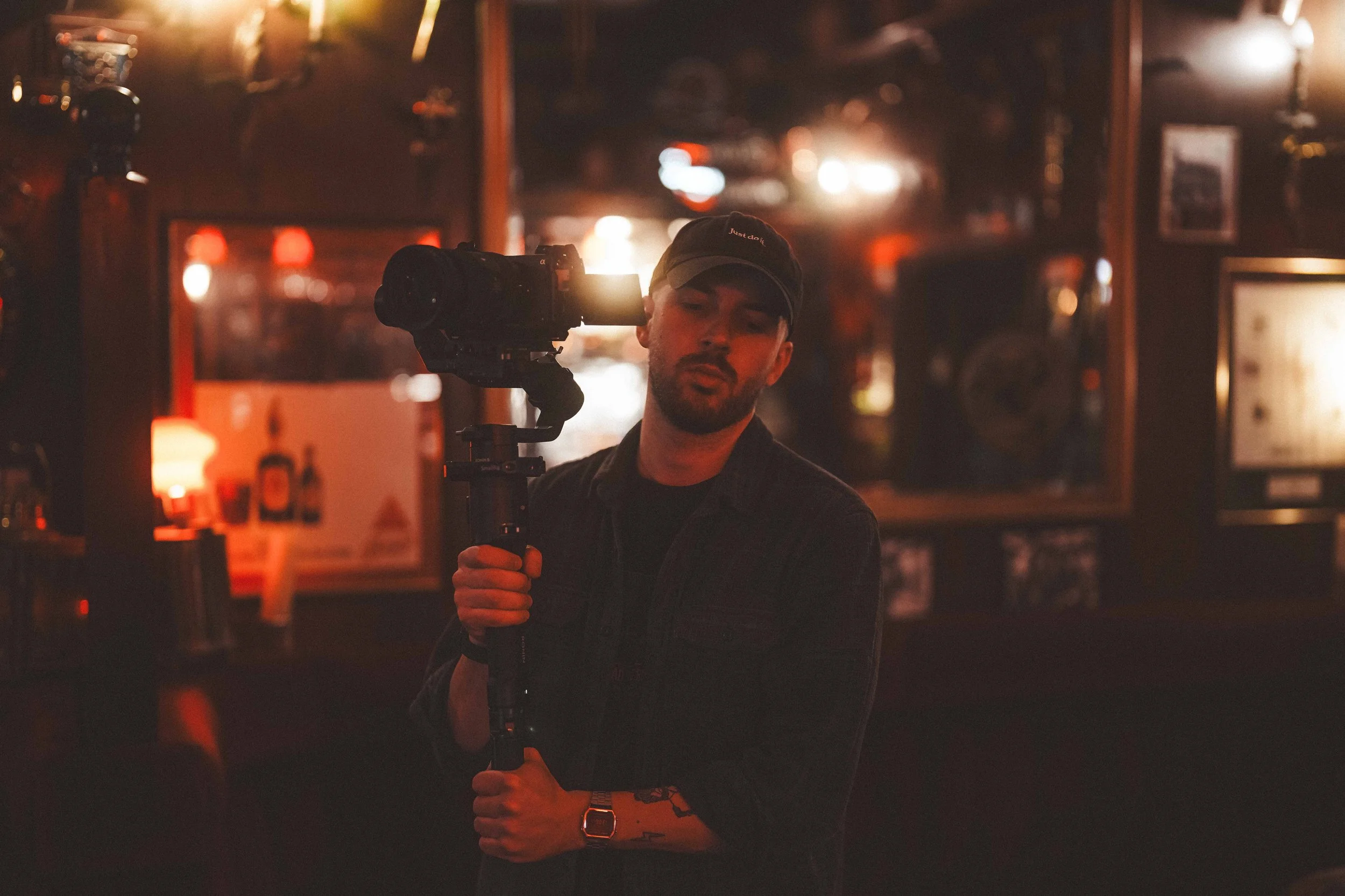 A man with a beard wearing a baseball cap and black shirt, holding a camera on a stabilizer in a dimly lit bar or club with warm reddish lights in the background.