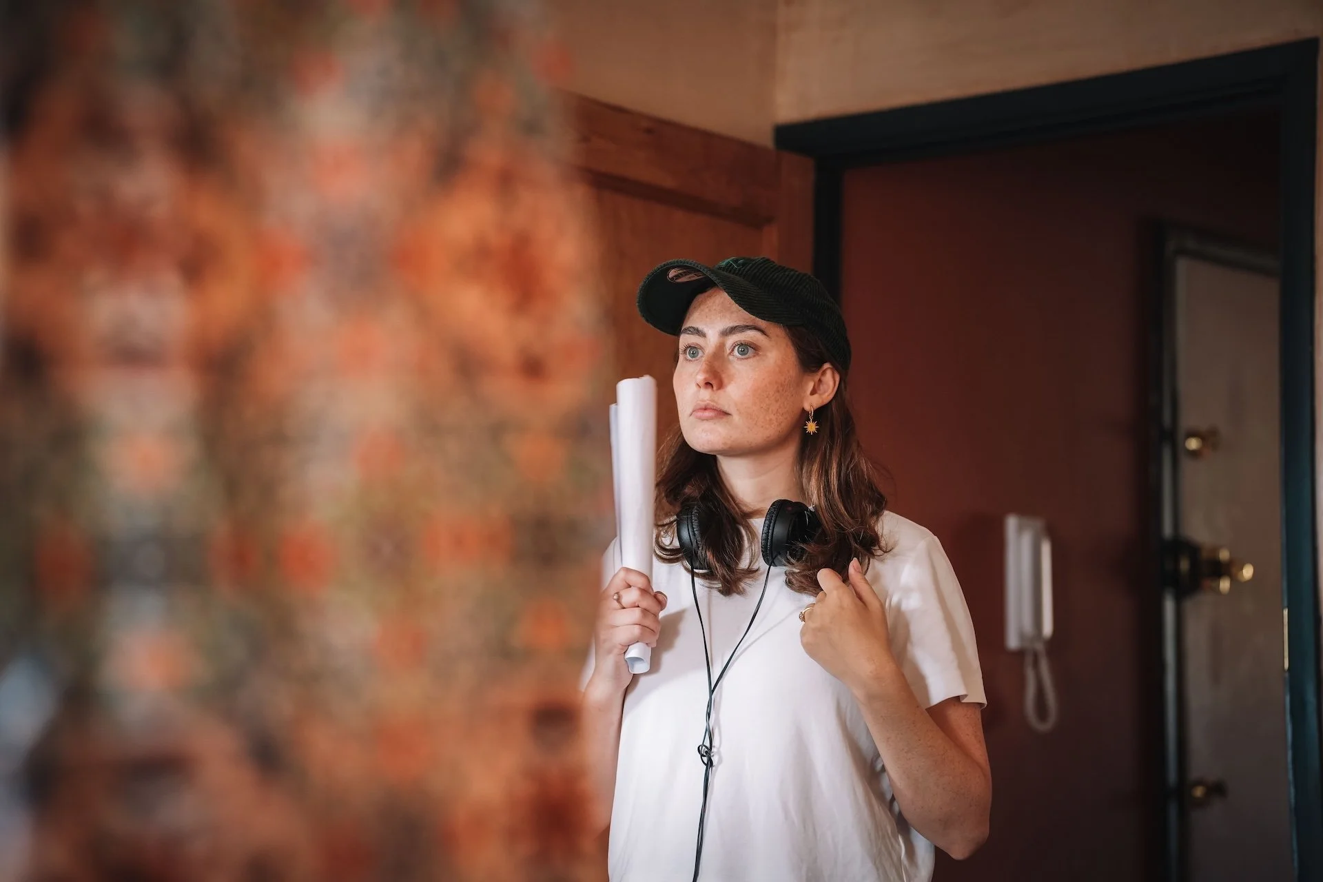 A woman with brown hair, wearing a black cap, white t-shirt, gold star-shaped earrings, holding a rolled-up piece of paper in front of a colorful blurred background. She has headphones around her neck and appears to be looking at something off-camera