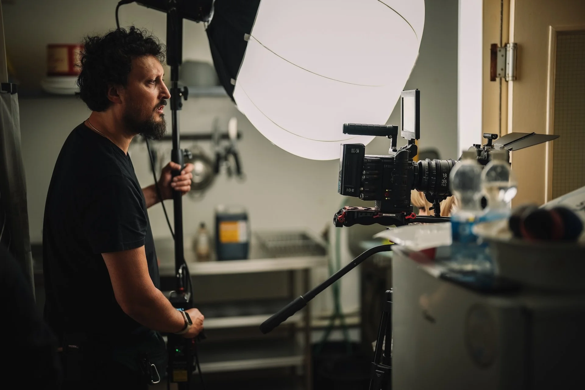 A man with curly hair and a beard operating a camera setup in a room with a large light diffuser overhead.