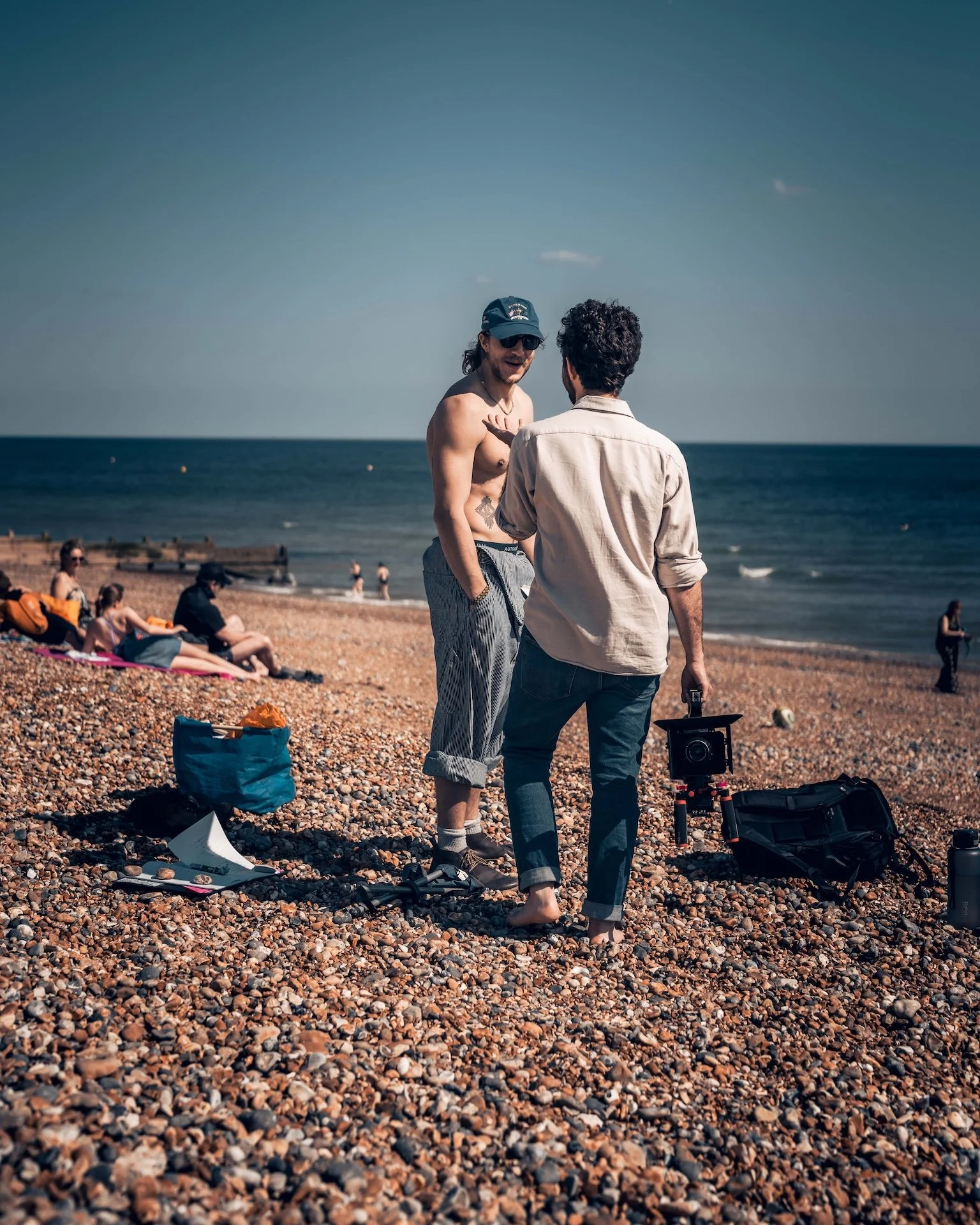 Two men on a pebbled beach, one shirtless wearing sunglasses and a cap, the other in a beige shirt with dark curly hair, filming with a camera, with people relaxing on the beach and the ocean in the background.