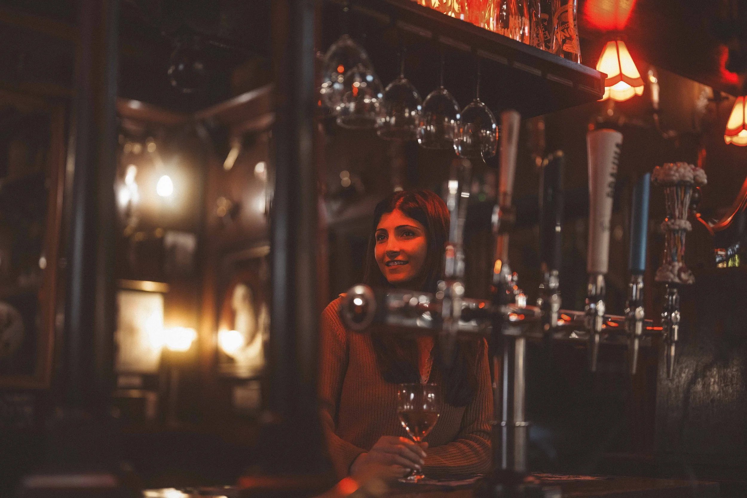 A woman with long dark hair smiling while holding a glass of champagne in a dimly lit bar, with hanging wine glasses and bar equipment visible around her.