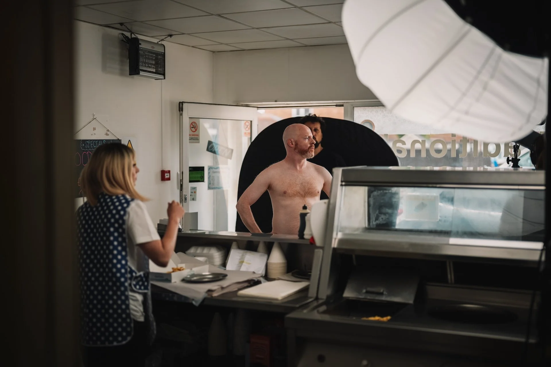 A shirtless man with a bald head and a beard standing behind a counter in a store, while a woman with blonde hair and a polka dot blouse stands in front of him. The view is partly obstructed by a large white umbrella-shaped light diffuser and a black