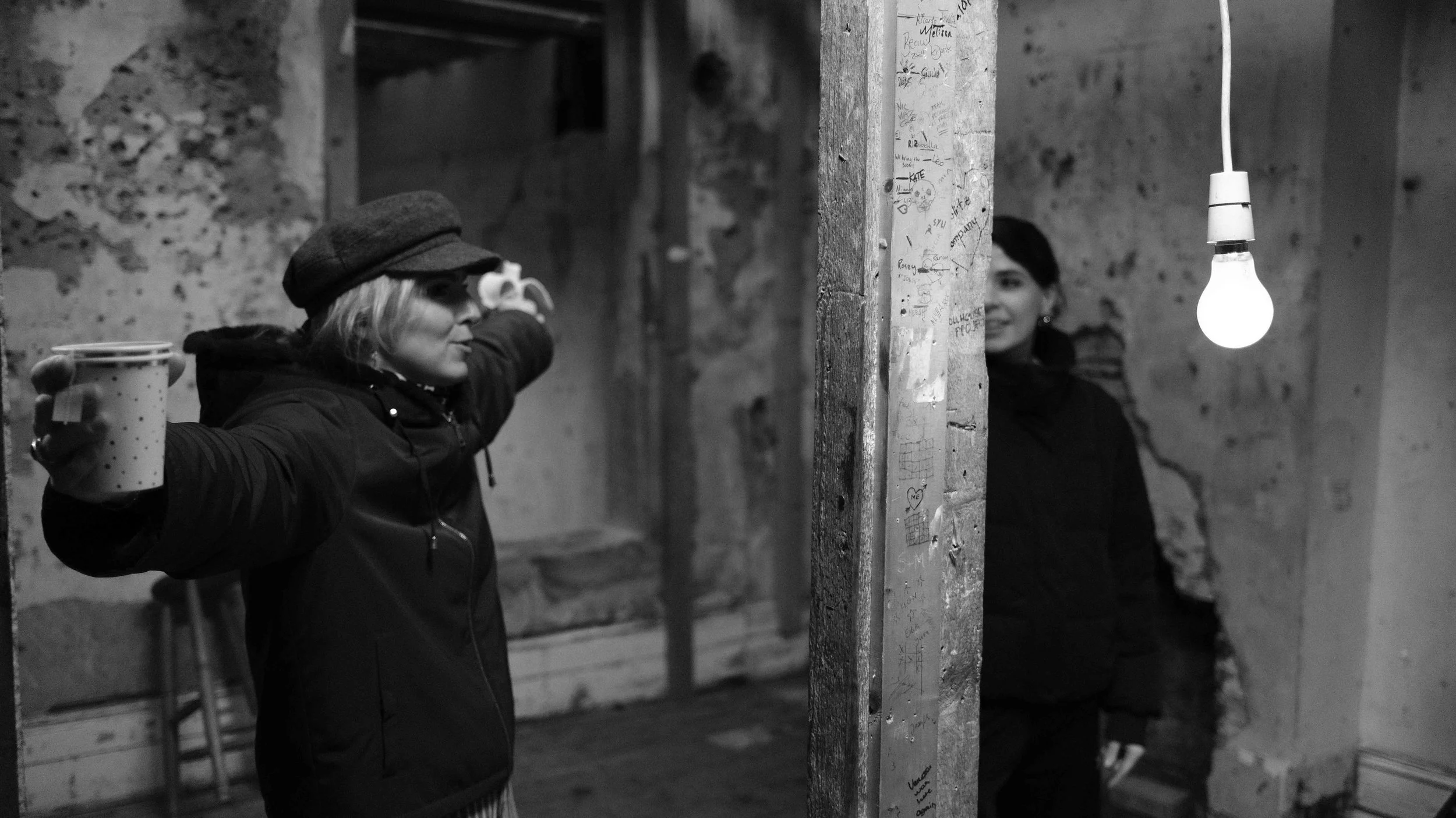 Two women standing in an old, rough brick and plaster room divided by a partition wall. The woman on the left is holding a polka-dotted mug and pointing, wearing a dark jacket and a hat. The woman on the right is smiling, dressed in dark clothing, an