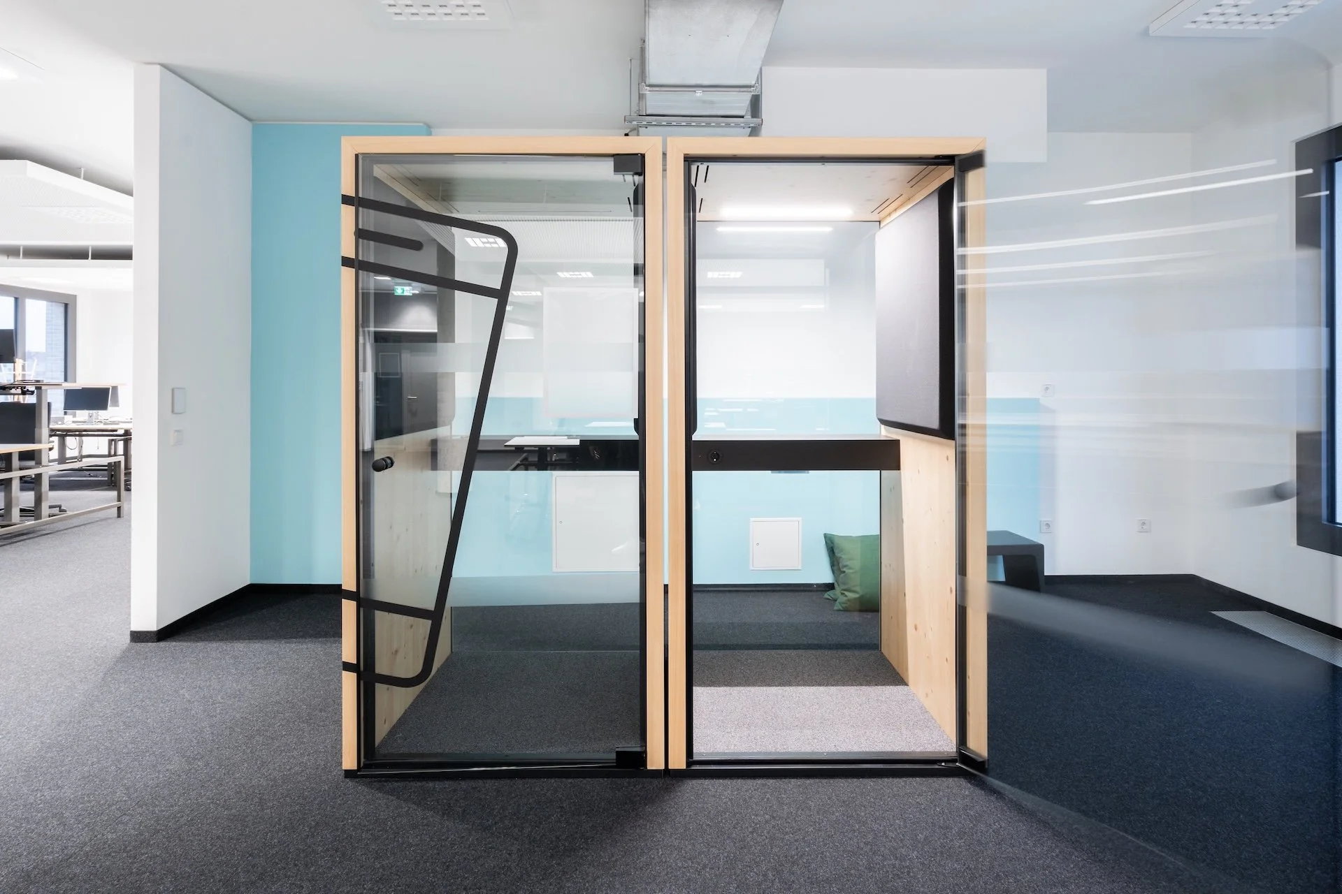 Modern glass and wooden office pod enclosed with glass walls and door, featuring black framing, a built-in desk, and a green cushion on the floor, situated in a contemporary office space.