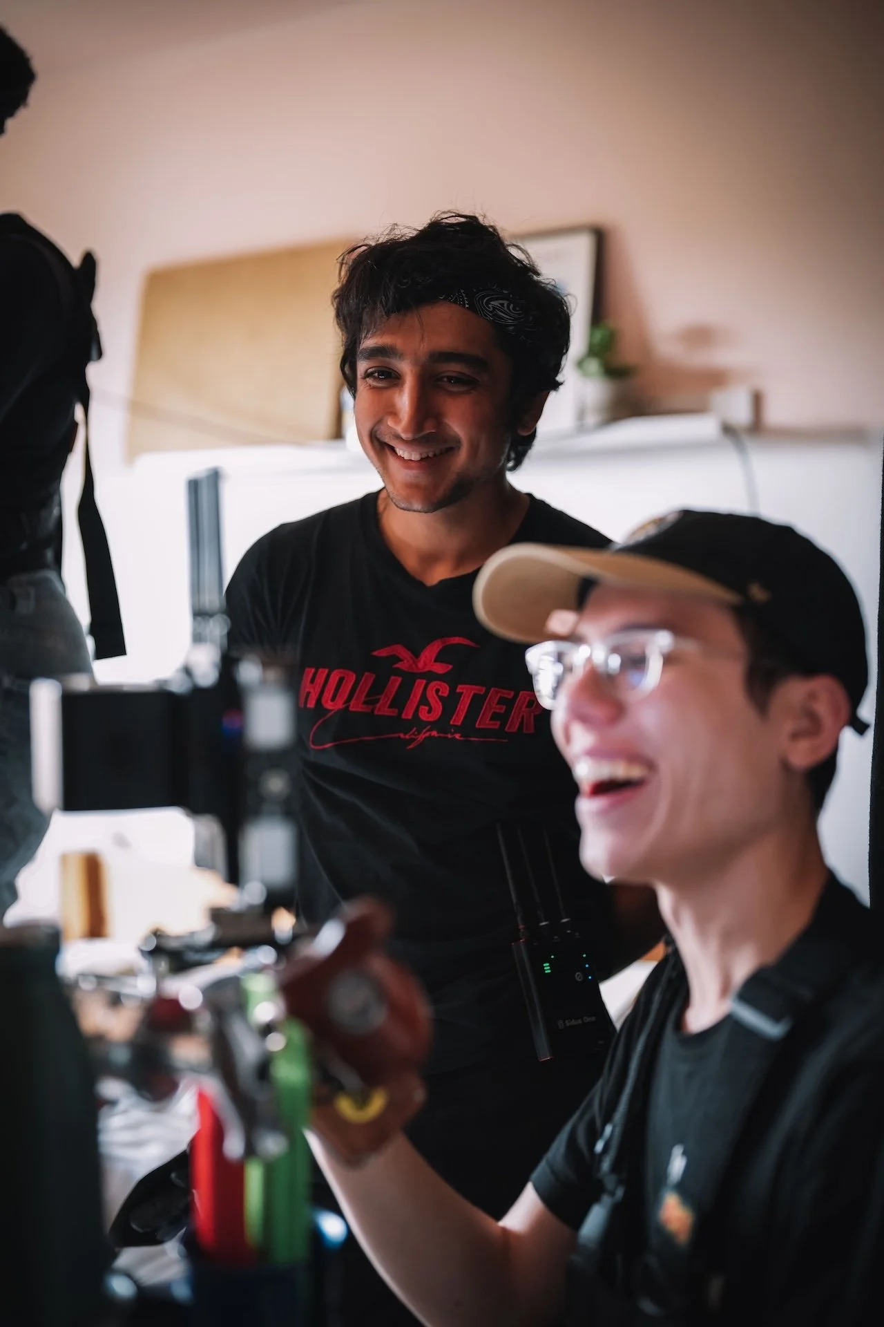 Two young men with dark hair smiling and working on a camera or filming equipment in a well-lit indoor space.