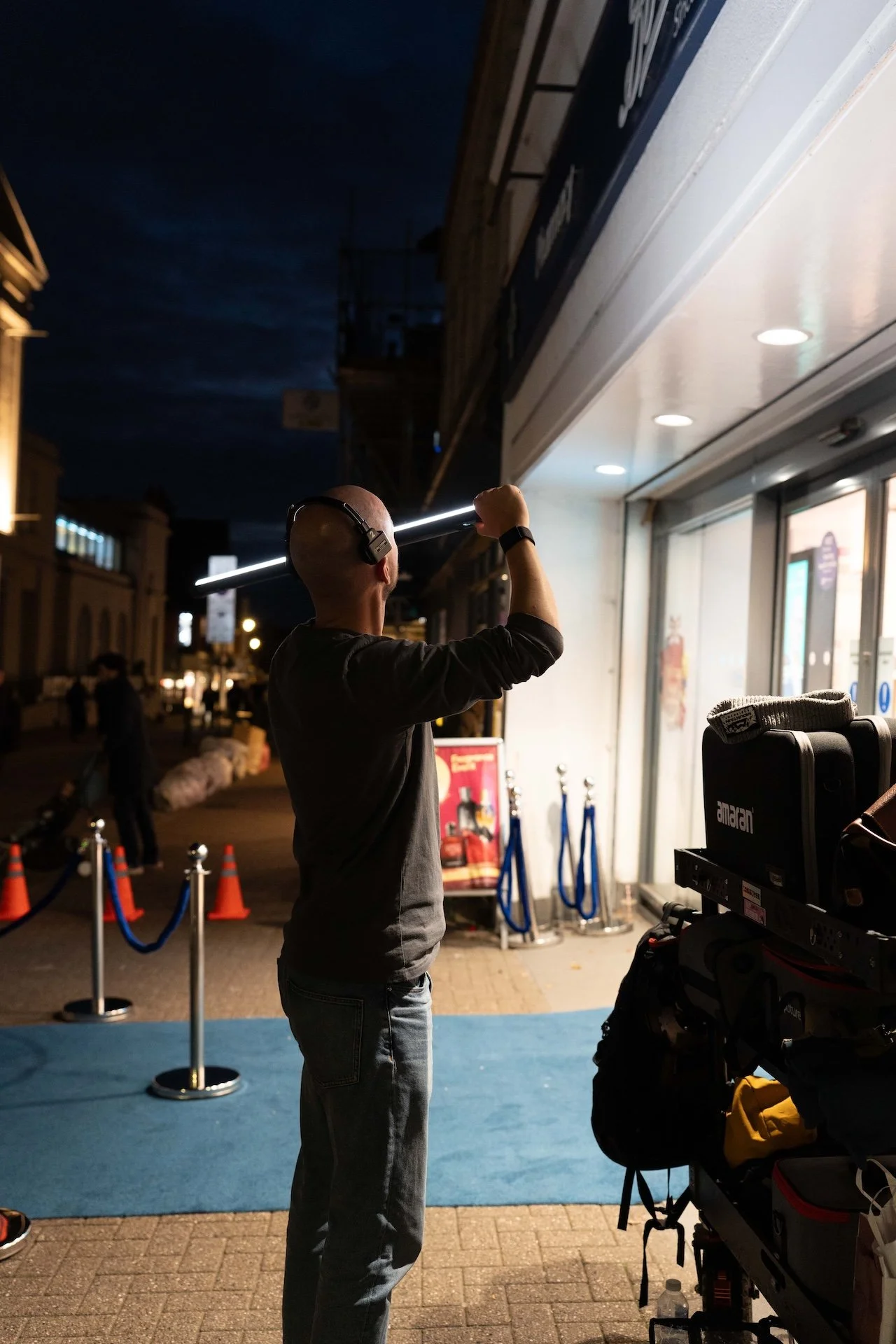 A person wearing headphones and a smartwatch is standing outside at night, holding a long light stick near their eye. The background features a city street with buildings, streetlights, and a line of orange cones and blue ropes.