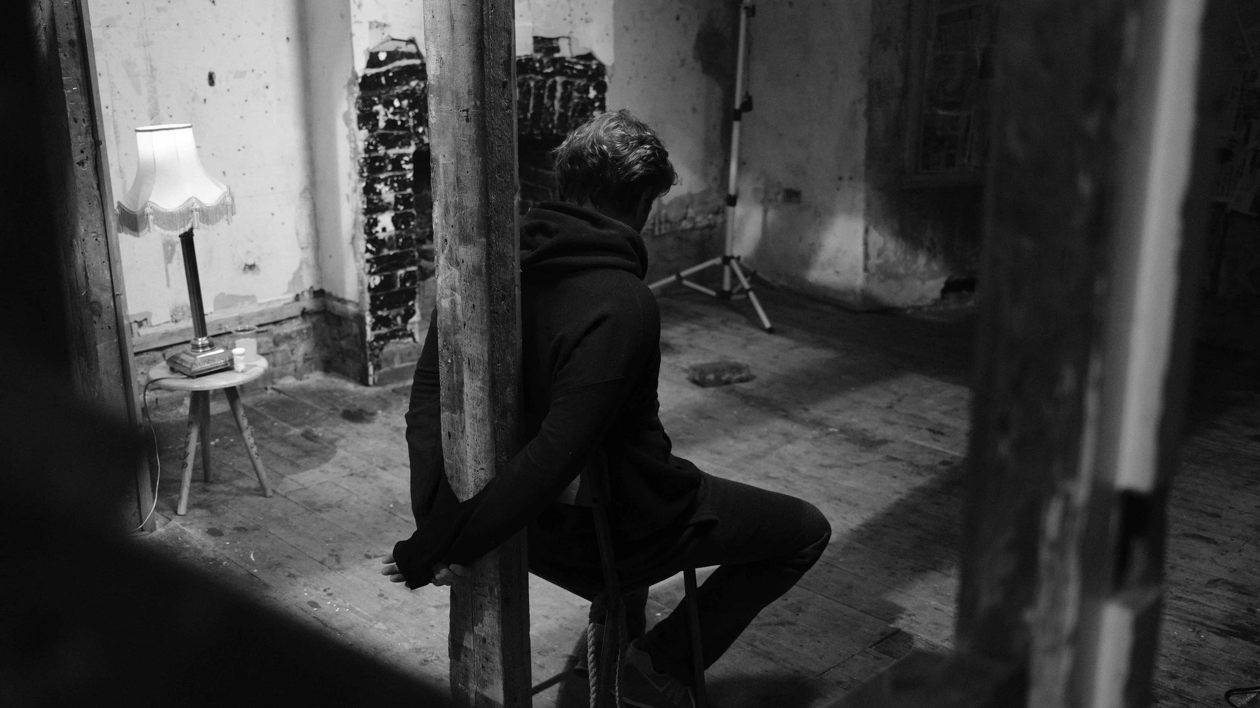 A person sitting on a chair with hands behind their back, in a dimly lit, rustic room with exposed brick and wooden beams, next to a small table with a lamp and books.
