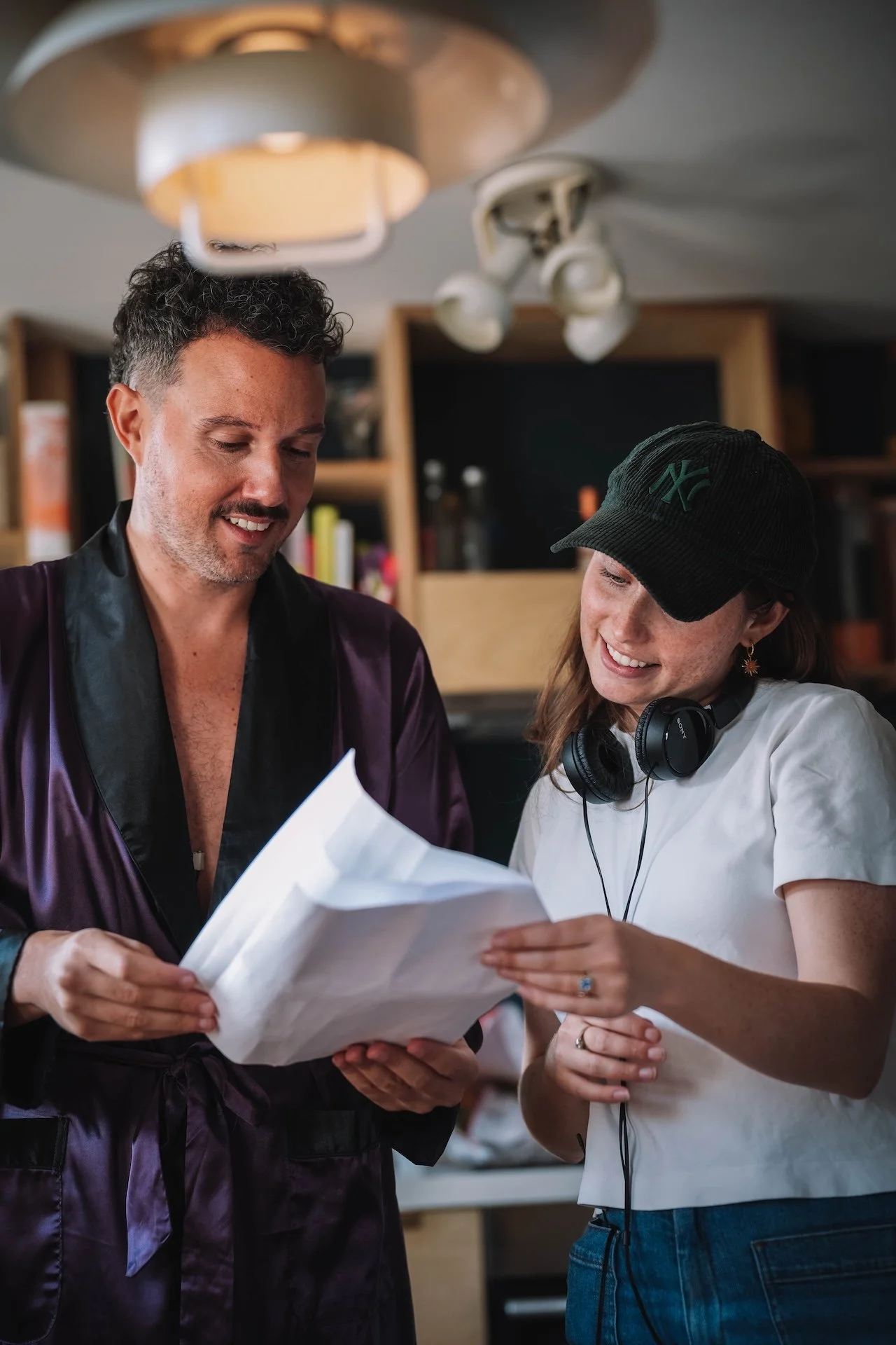 Two women looking at papers and smiling indoors, one wearing a black cap, headphones around neck, and white shirt, the other in a satin purple robe with black lapel.