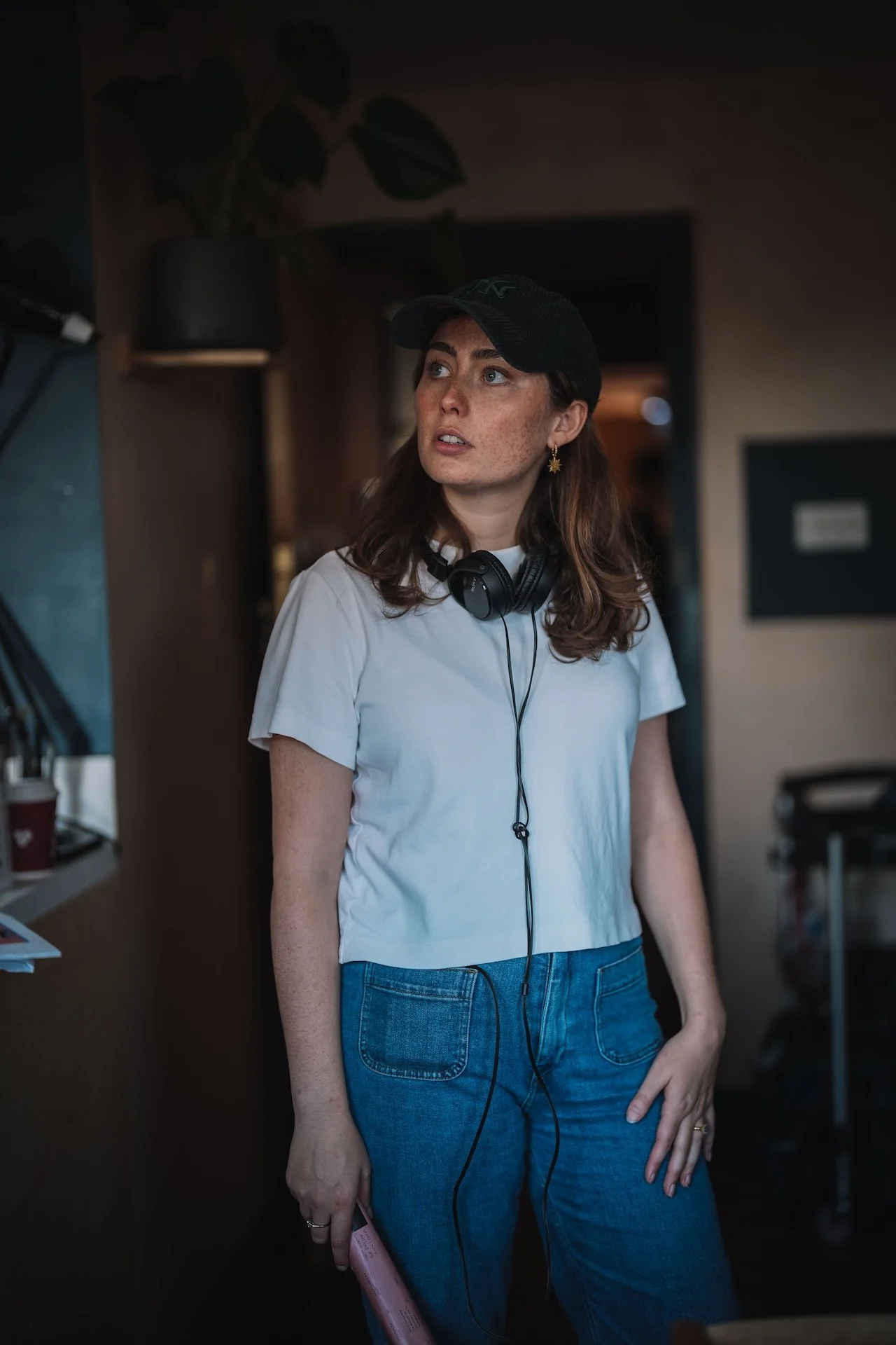 A woman with shoulder-length brown hair, freckles, and light blue eyes is standing indoors. She is wearing a black cap, gold earrings, a white t-shirt, and blue jeans. She has headphones around her neck and is holding a pink object in her right hand.