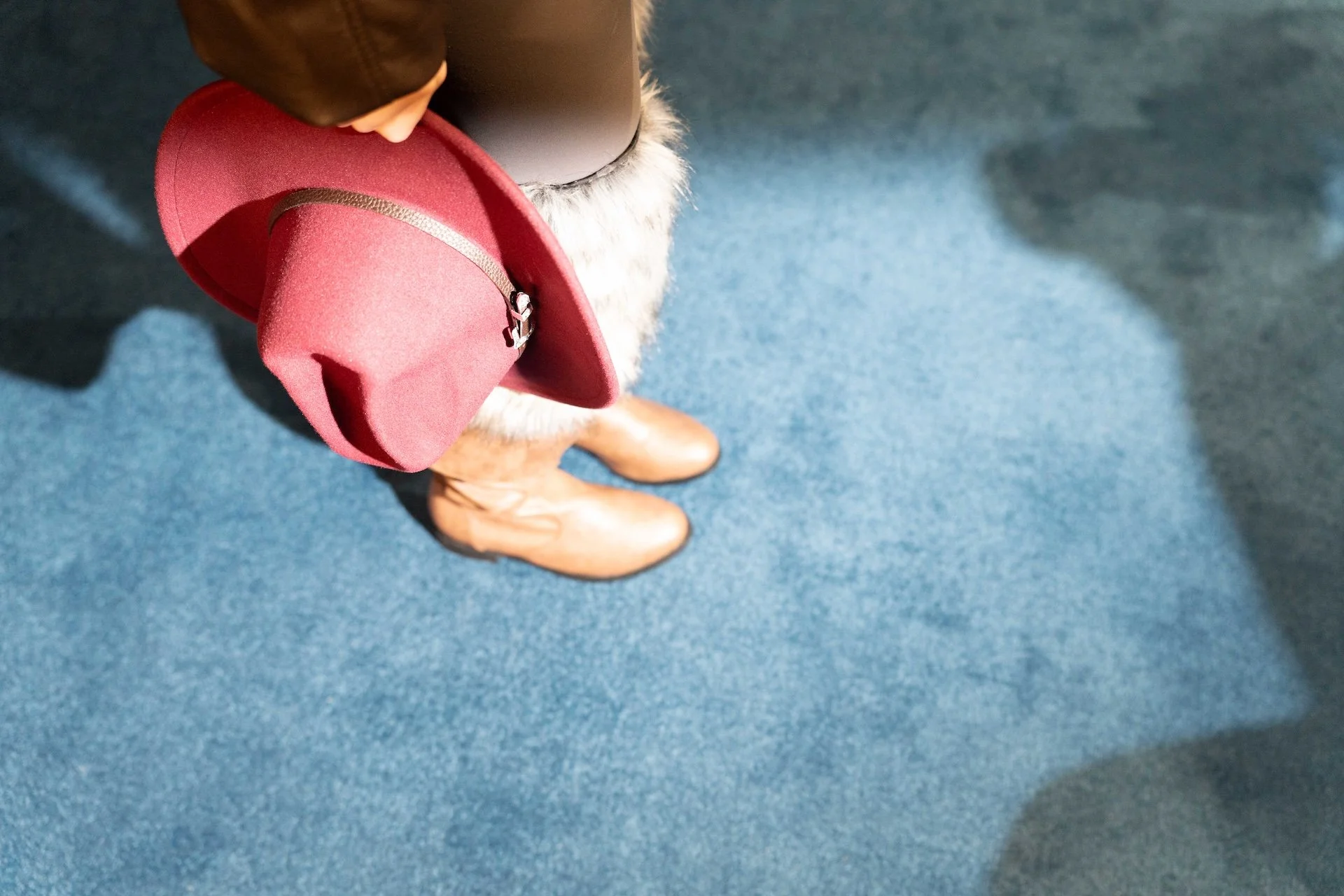 The image shows a top-down view of a person standing on a blue carpeted floor. The person is wearing beige cowboy boots, a brown coat, and dark pants. They are holding a pink wide-brimmed hat with a black band.