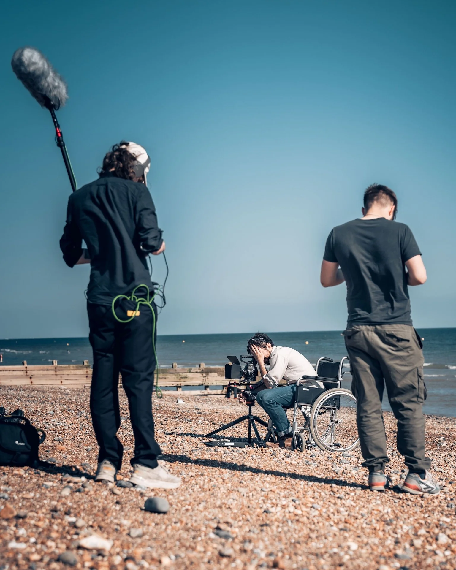 Film crew filming on a beach with a camera operator in a wheelchair, crew members surrounding him, and the ocean in the background.