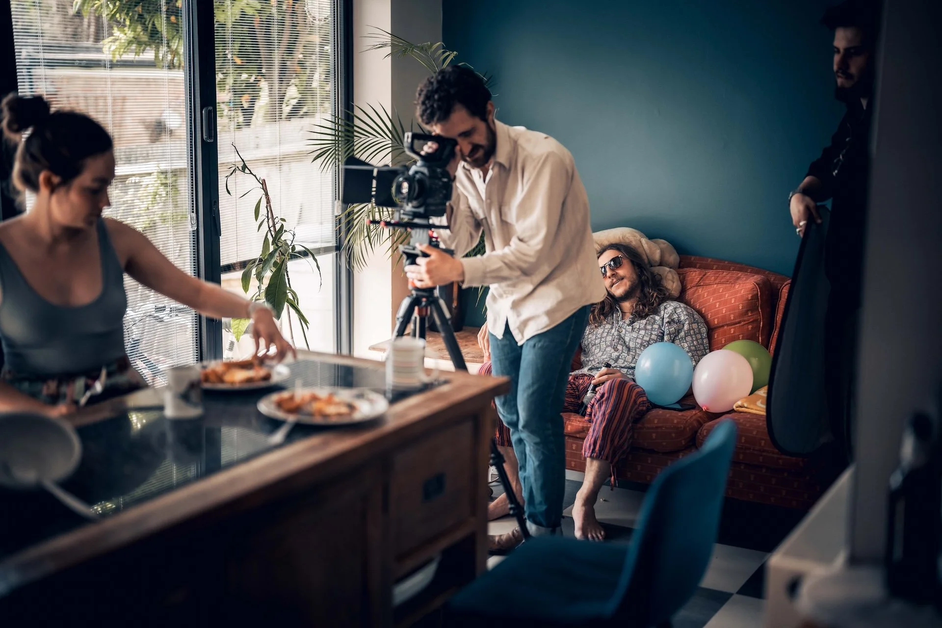 People celebrating with balloons and food during a video shoot in a living room.