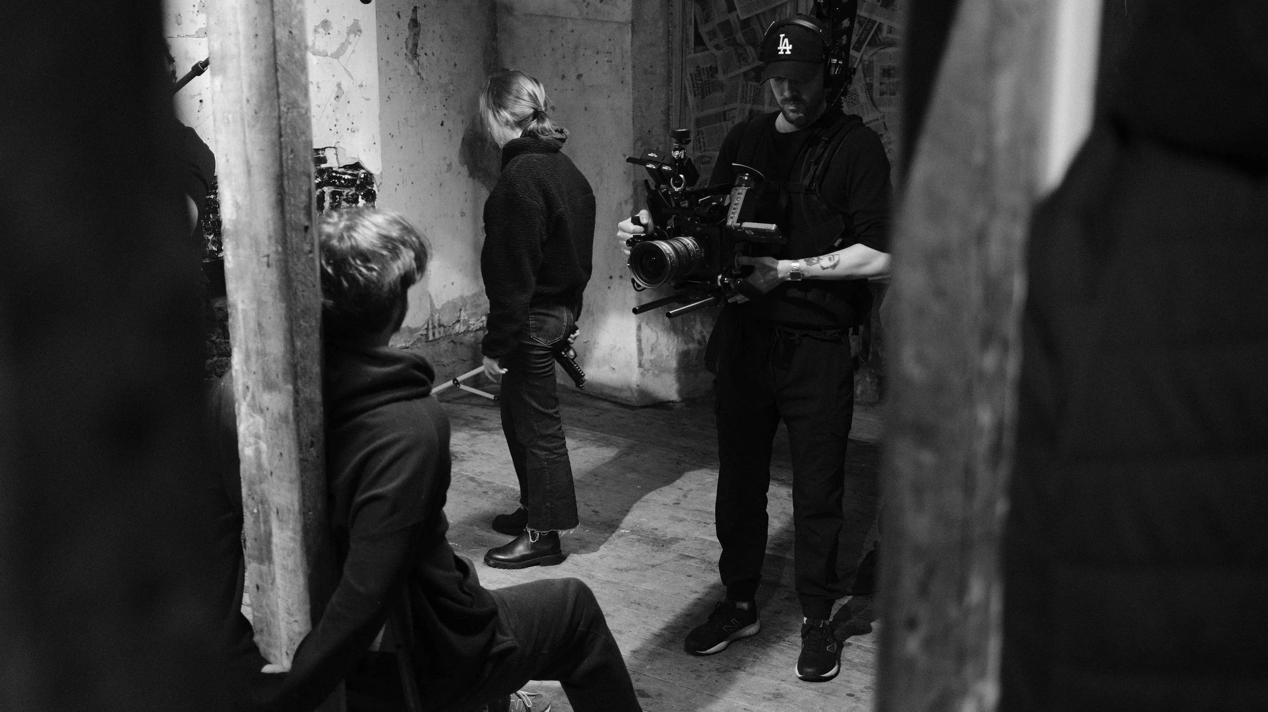 Black and white photo of a film crew filming indoors. A man wearing a Los Angeles Dodgers cap holds a camera, while two women and a man sit or stand nearby. Part of the scene is obscured by two vertical objects on the sides.