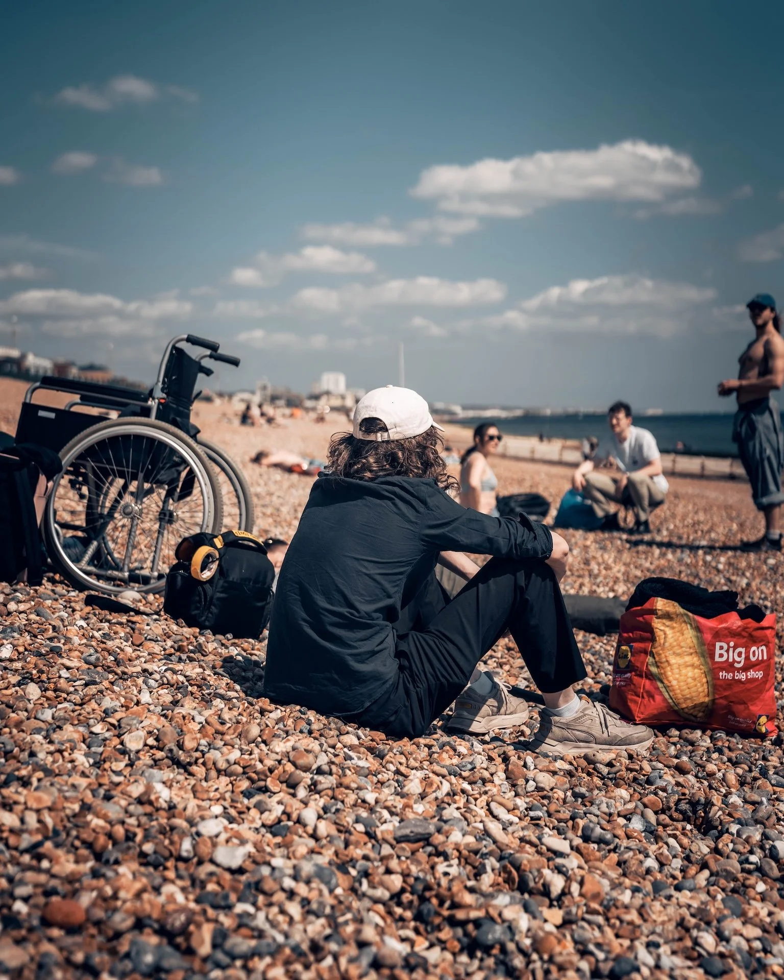 A person sitting on a pebble beach with their back facing the camera. They are wearing a white cap and black clothing, with a colorful bag beside them that has the text "Big on the big shop." In the background, there are several people, a wheelchair,