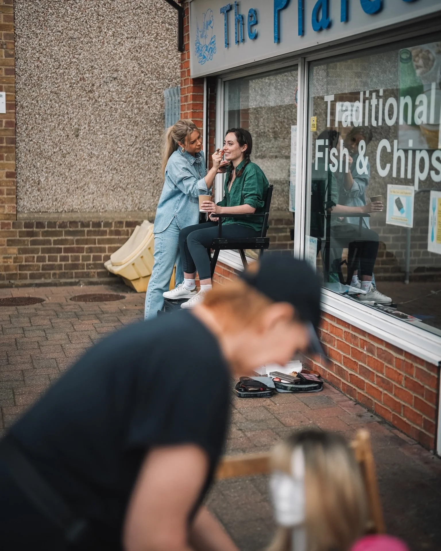 Two women outside a fish and chips shop, with one woman sitting on a chair and the other woman applying makeup or lipstick to her face. The shop window has text that reads 'Traditional Fish & Chips'. The scene is viewed through a blurred foreground, 