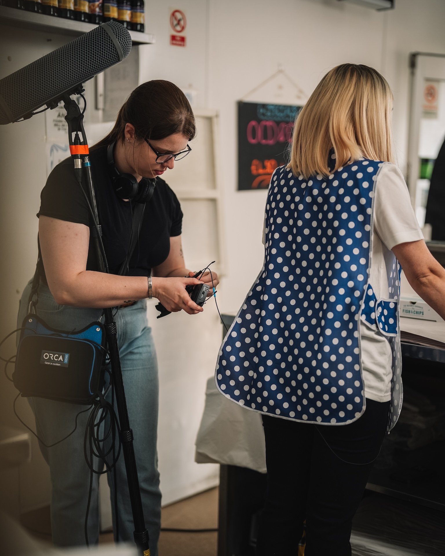 A person with glasses and headphones standing next to a scooter while adjusting some equipment, with another woman in a polka dot vest standing nearby in an indoor setting.