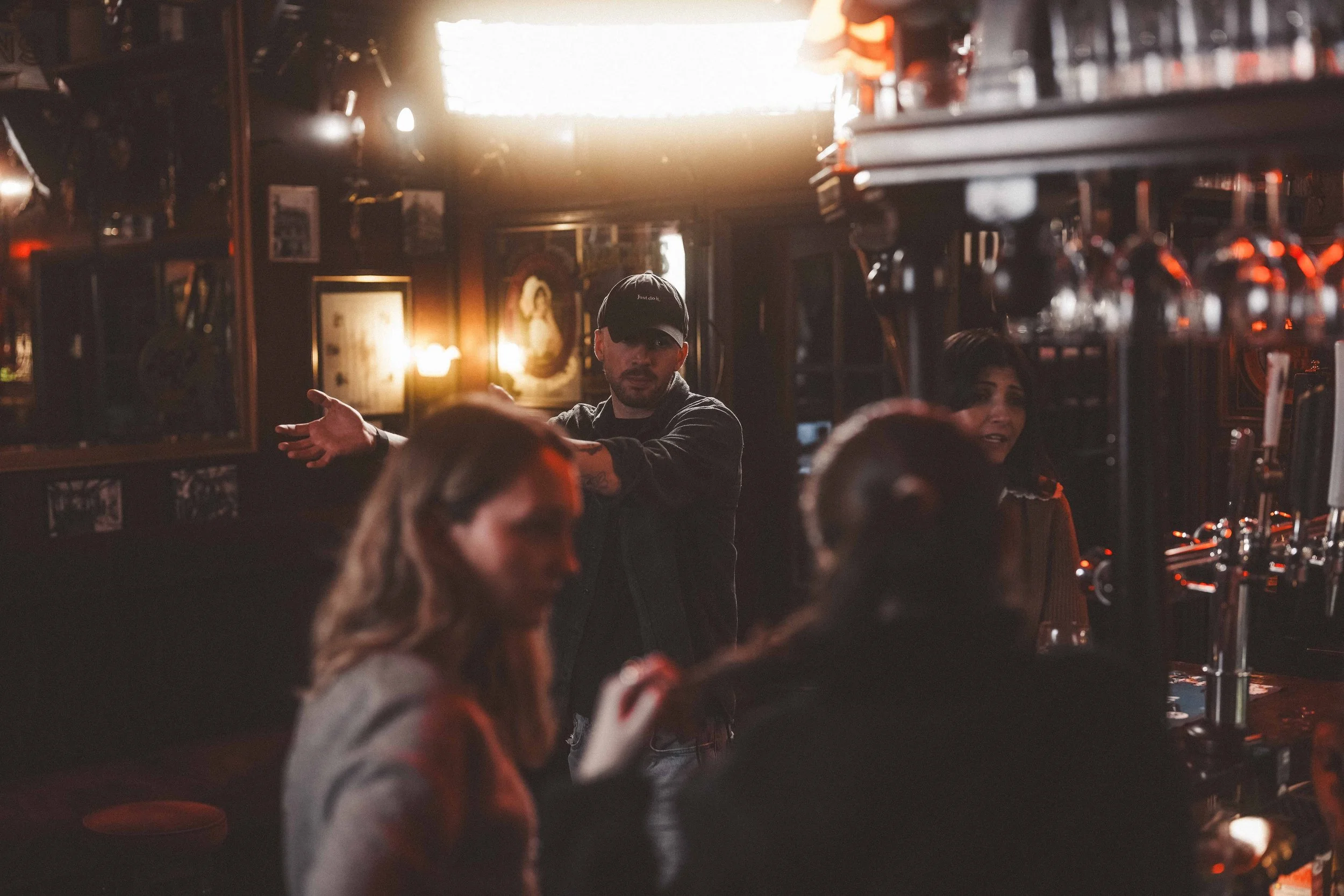 People inside a bar with dim lighting, one person in a black cap gesturing, others socializing, shelves with bottles and bar equipment visible.