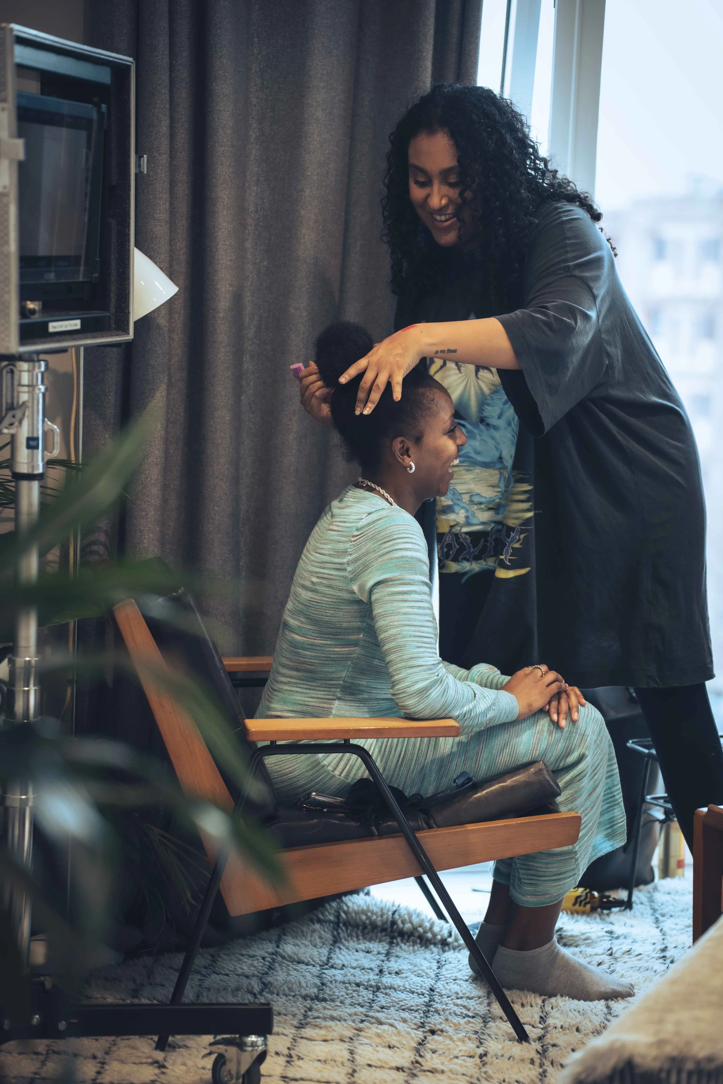 A woman sitting on a chair smiling while getting her hair styled by another woman standing next to her in a cozy indoor space with large windows and dark curtains.