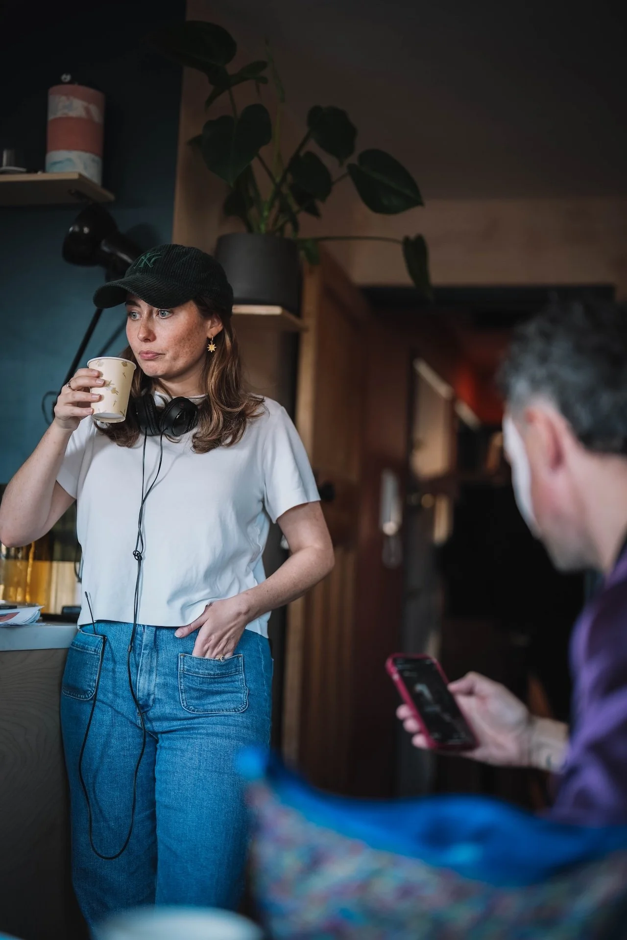 A woman wearing a black cap, white t-shirt, and blue jeans is standing in a cozy indoor space with wooden walls and a large potted plant. She is sipping from a disposable cup and has headphones around her neck. A man with glasses is sitting nearby, u