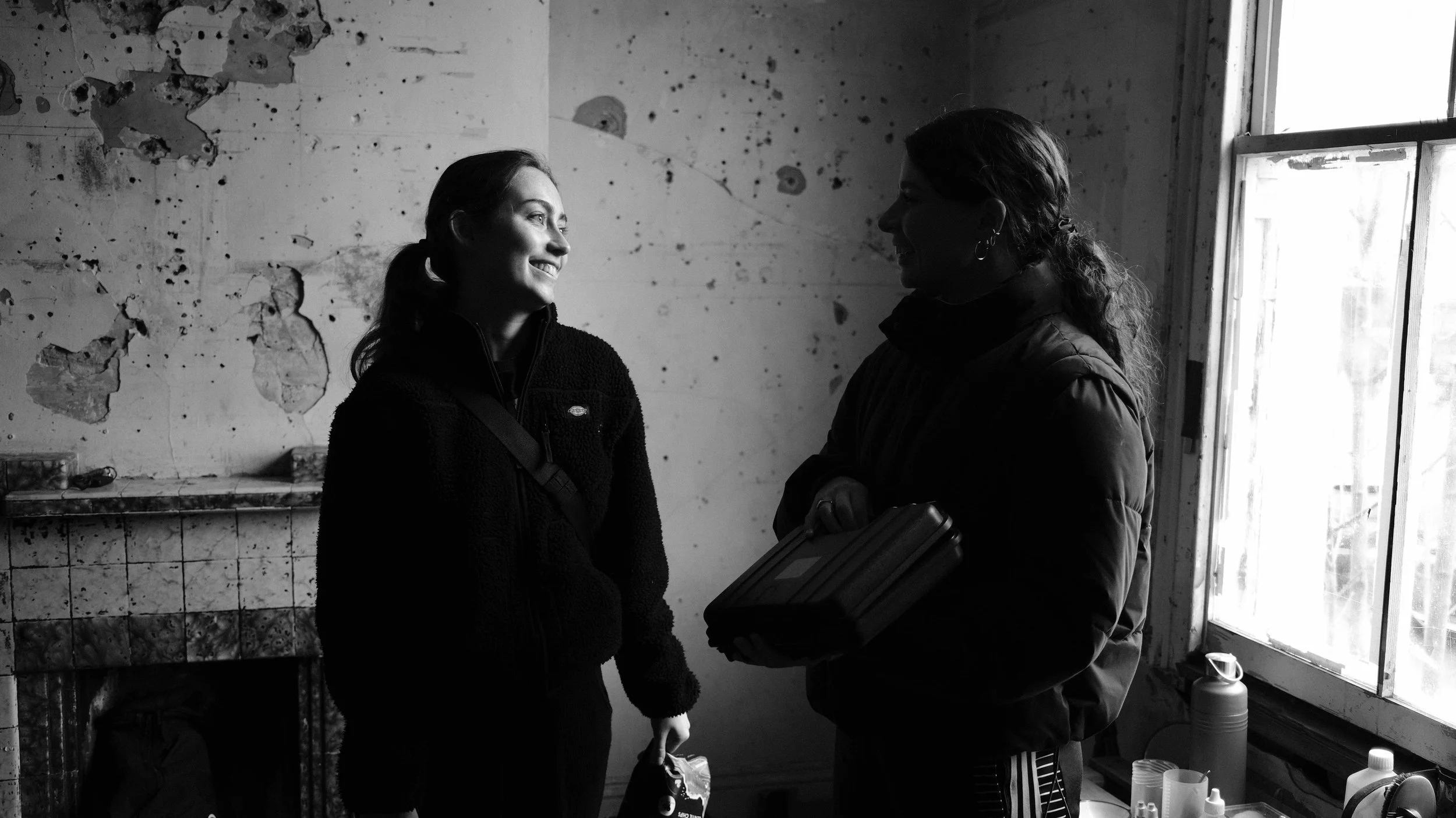 Two women smiling and talking inside a room with a window. One woman holds a bag, the other holds a closed laptop. The room has peeling paint and a cluttered table.