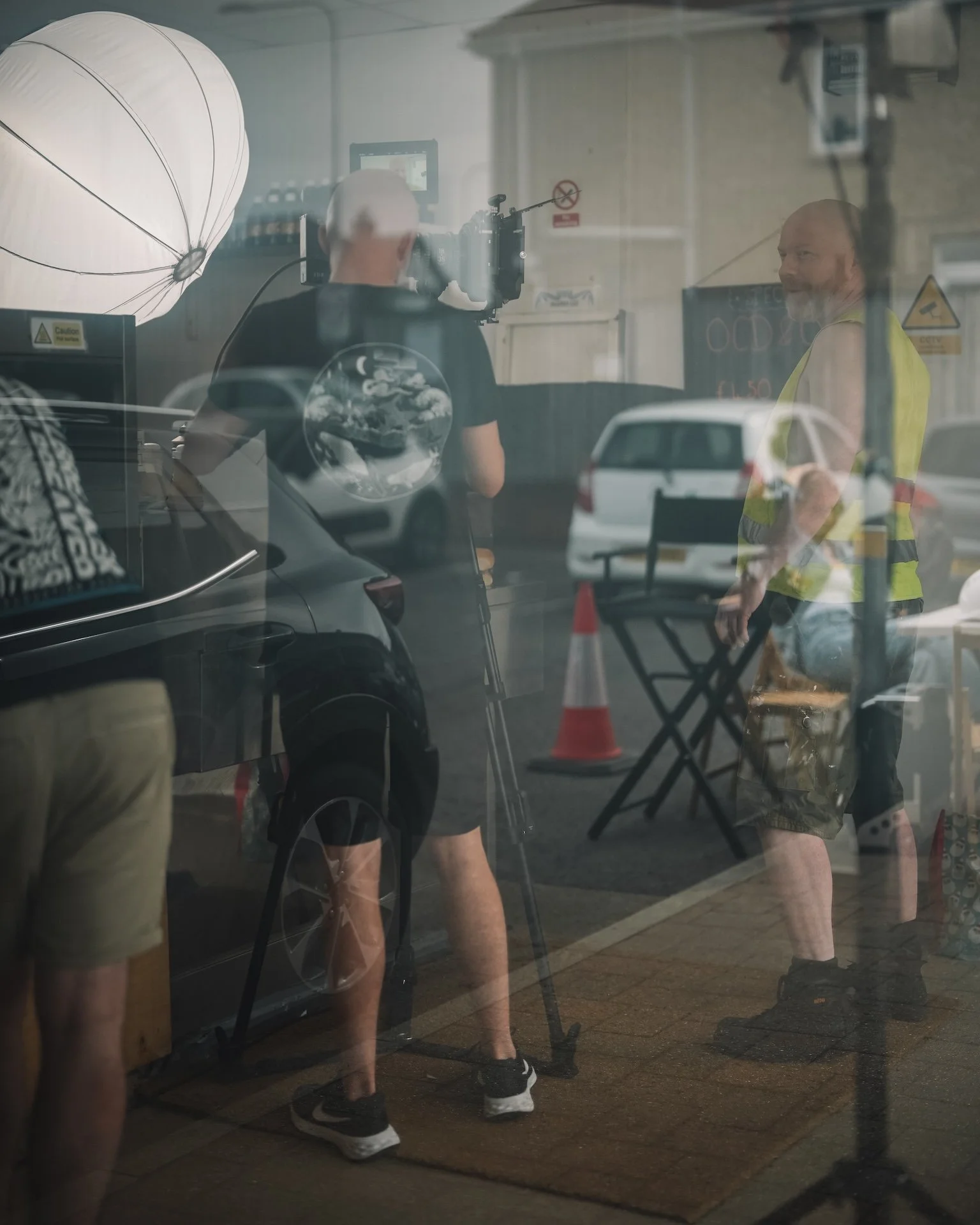 A reflection of two men standing outside a building with a camera, lighting equipment, and a chalkboard sign visible through the glass window. One man is wearing a black T-shirt and shorts, the other is wearing a yellow safety vest and camouflage sho