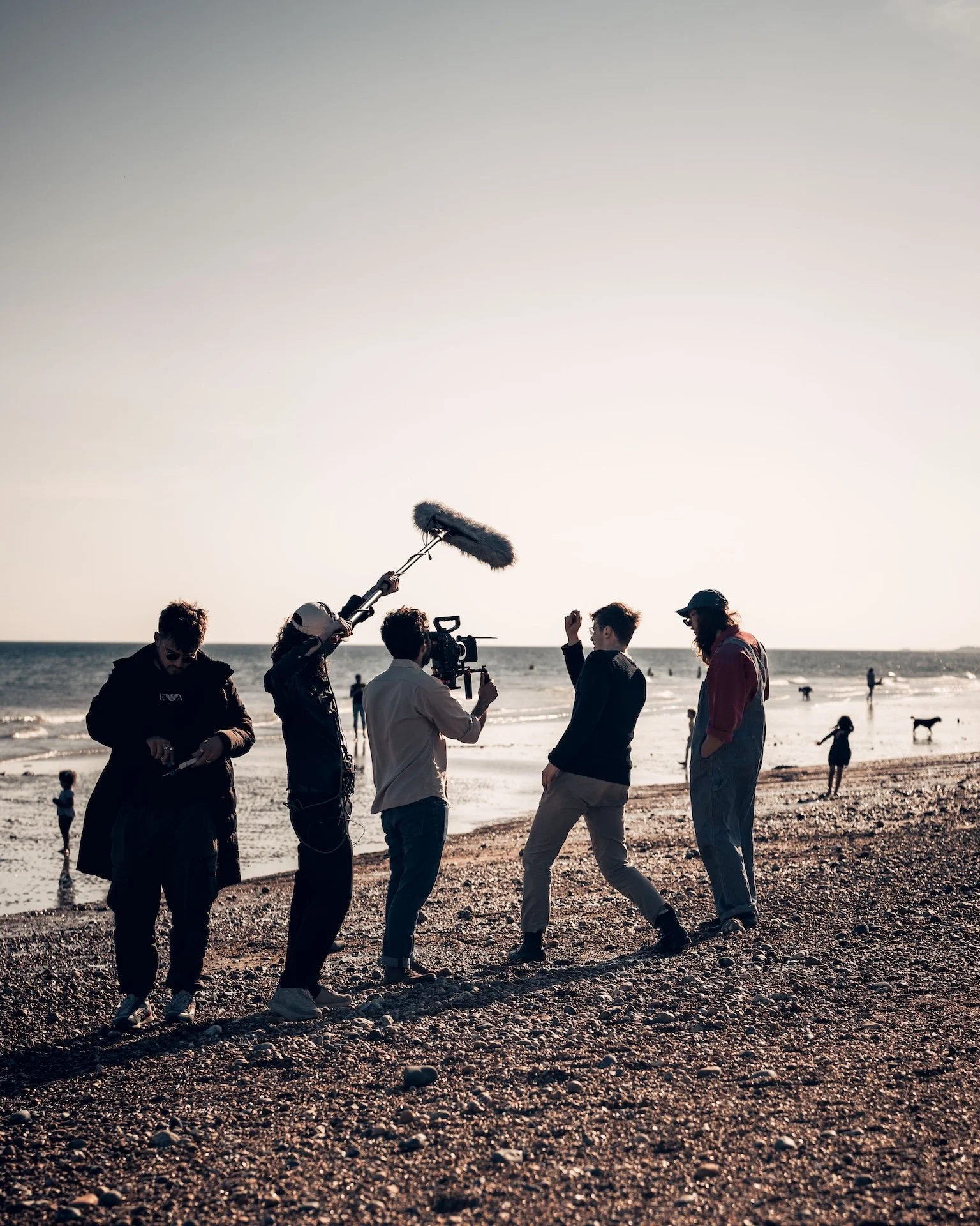 A film crew filming on a beach at sunset, with six people in the foreground and dogs and children in the background.