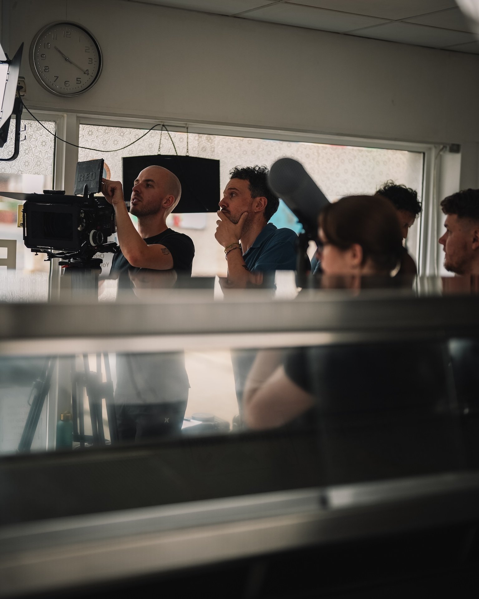 People sitting in a restaurant or cafe, with a man in a blue shirt and another man with a shaved head, sitting near a large window. A clock on the wall shows 4:59.