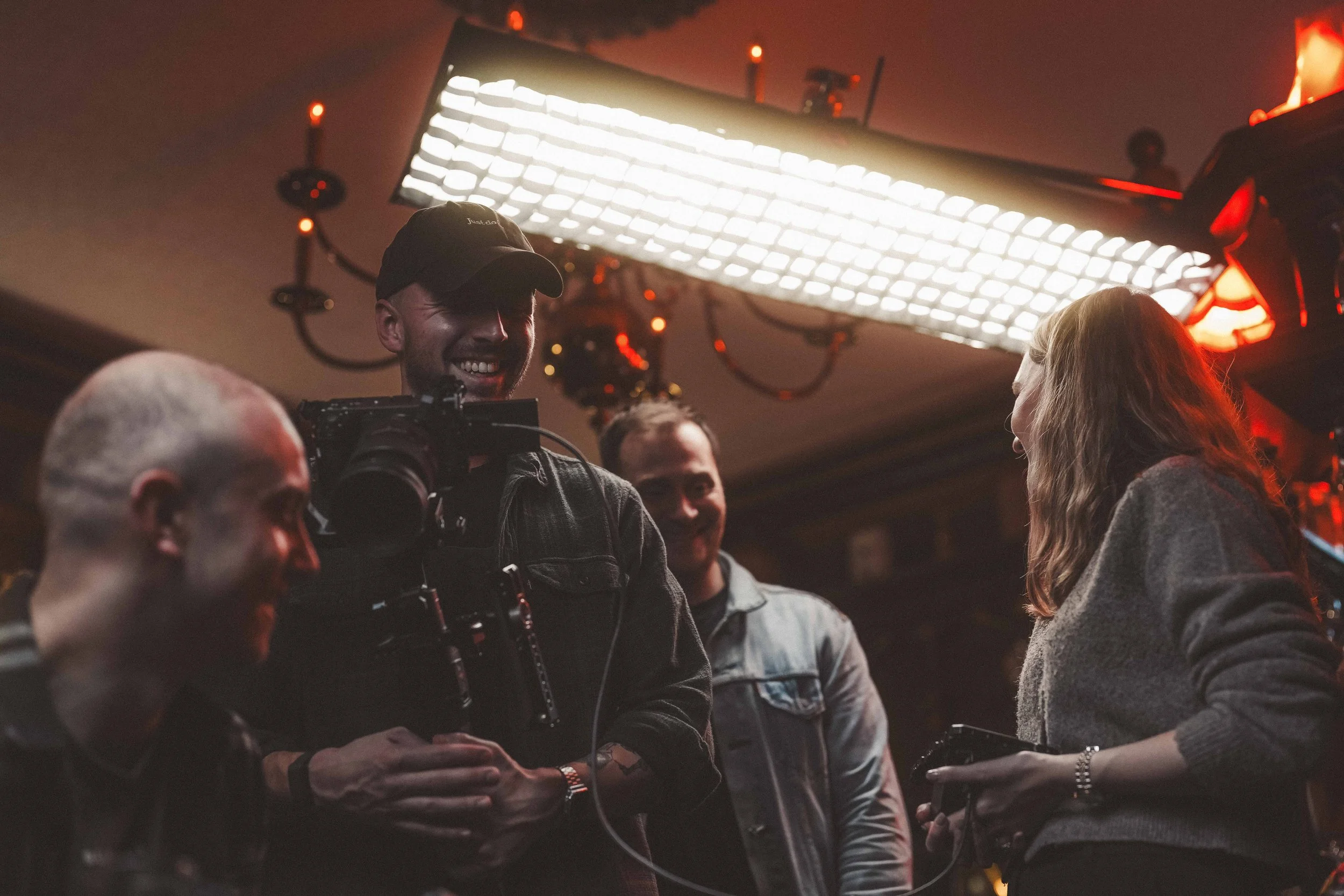 A group of people, including a man wearing a cap and a woman, are in a dimly lit room with red lighting, smiling and engaging in conversation. A large bright light panel is mounted on the ceiling.