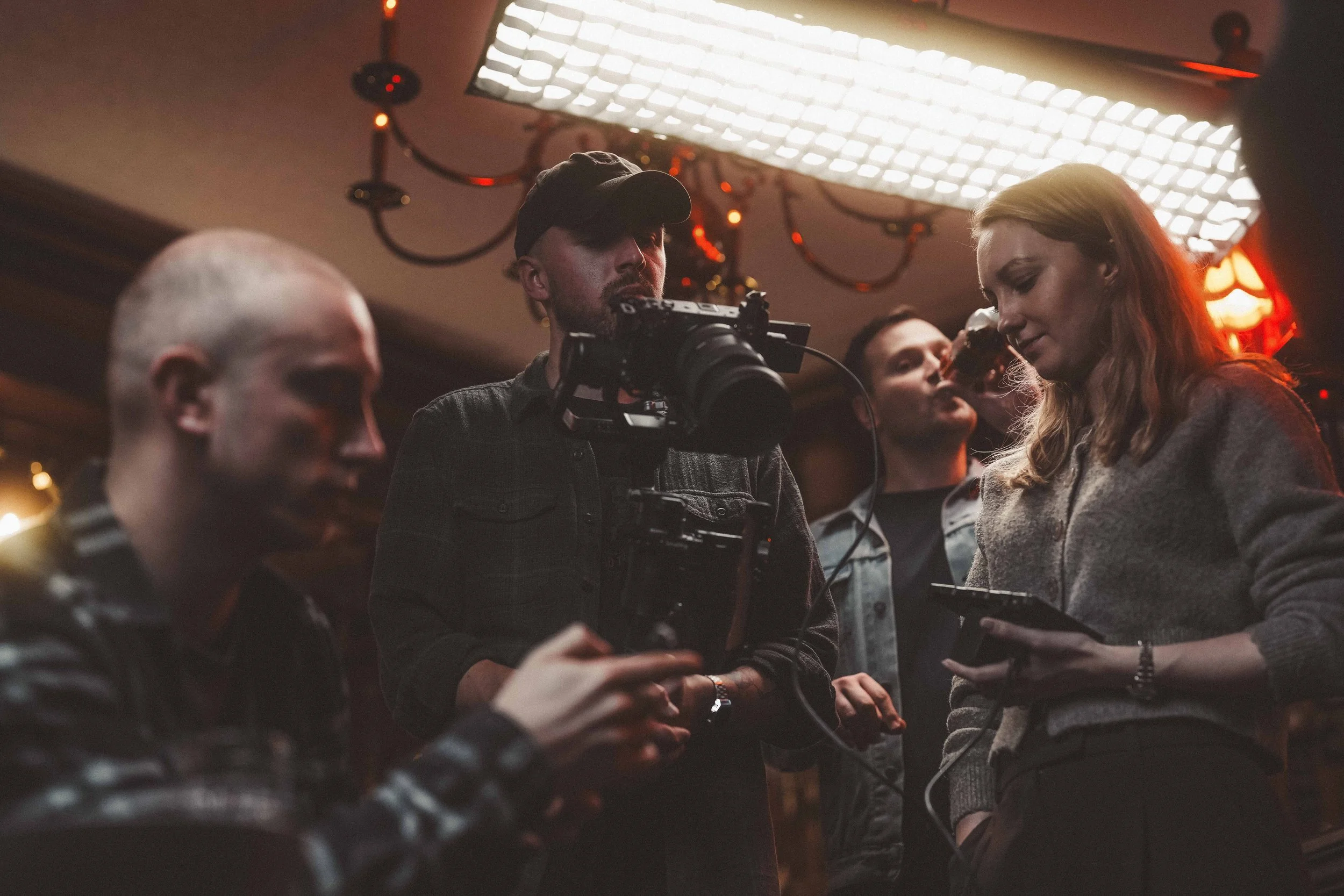 Group of people filming a scene in a dimly lit room with a large bright light overhead, focusing on a woman with long hair holding a drink, while a man with a camera and a woman stand nearby.