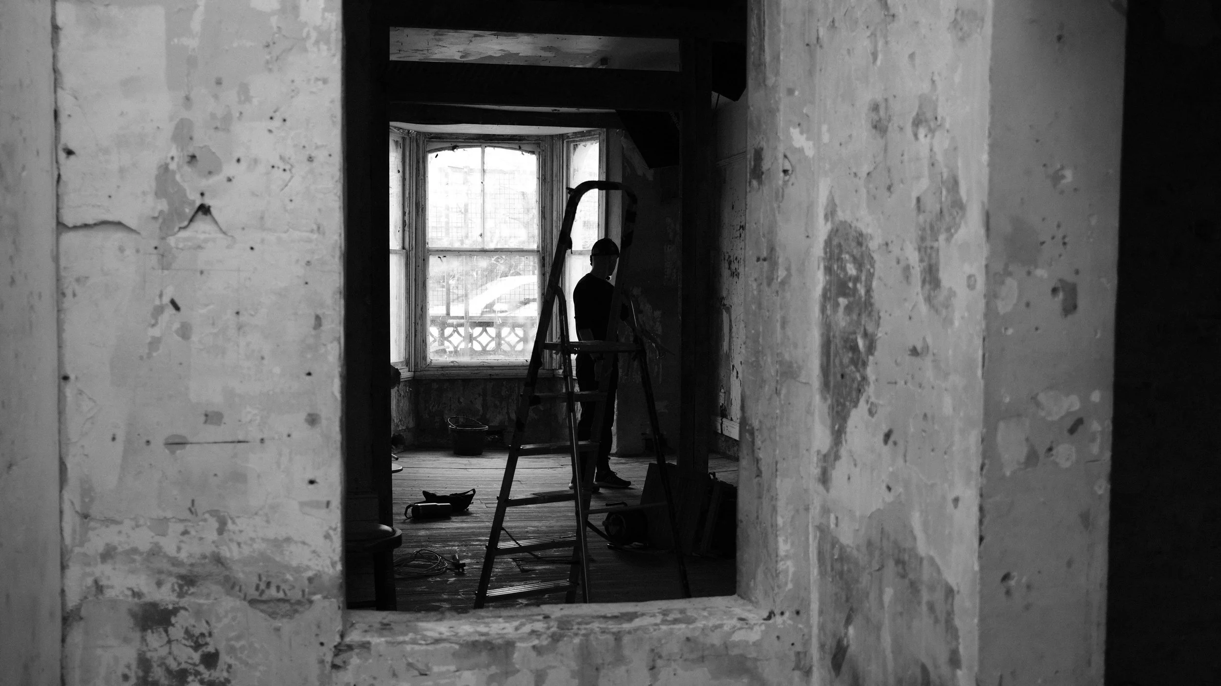 Black and white photo of a person standing on a ladder in a room with peeling paint walls and a large window in the background. The person is silhouetted against the light coming through the window.