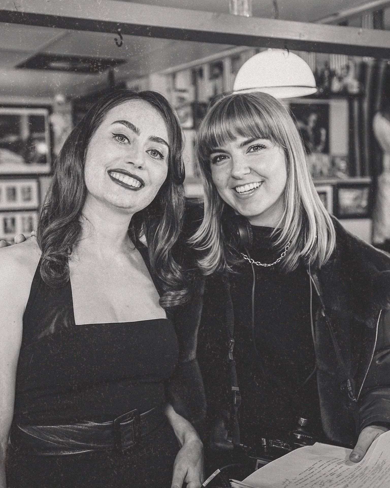 Two women are smiling and posing for a photo in an indoor setting with shelves and pictures in the background. One woman has dark hair and the other has blonde hair. The photo is in black and white.