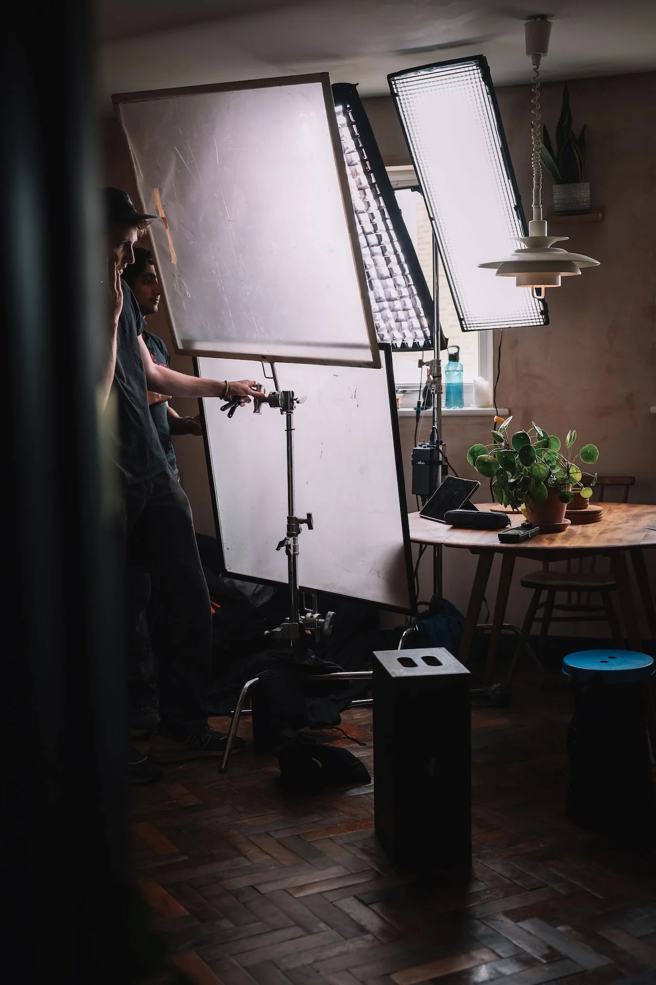 Photo of a filming or photography setup in a room with two people working behind a white backdrop, large lighting equipment, a wooden table with a potted plant, and a window with natural light.