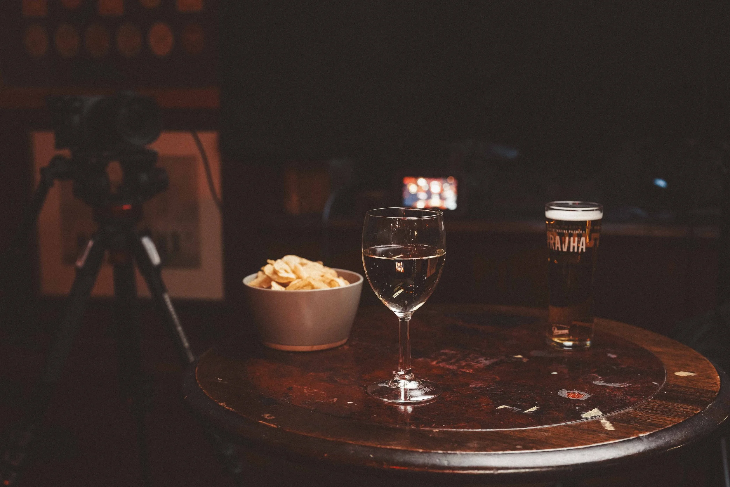 A table with a glass of white wine, a beer glass, a bowl of potato chips, and a camera on a tripod in a dimly lit room.