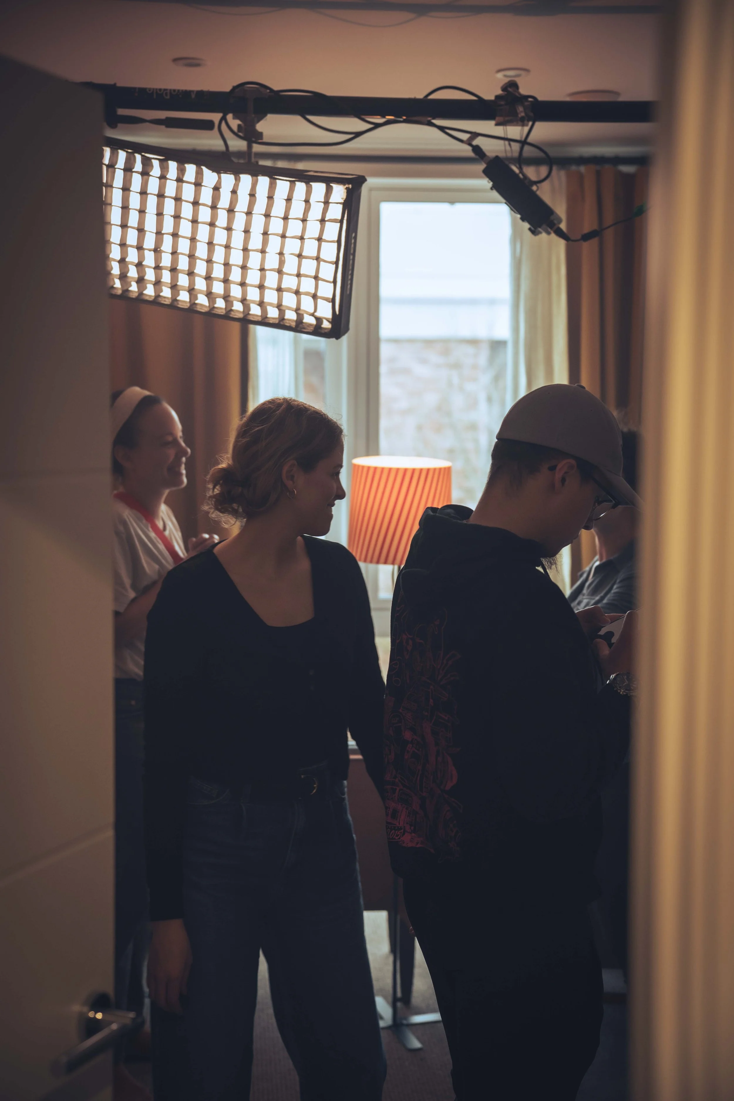 A group of people in a room, some smiling and some engaged with their phones, with a soft warm lighting, a window in the background, and a large professional light on a stand overhead.