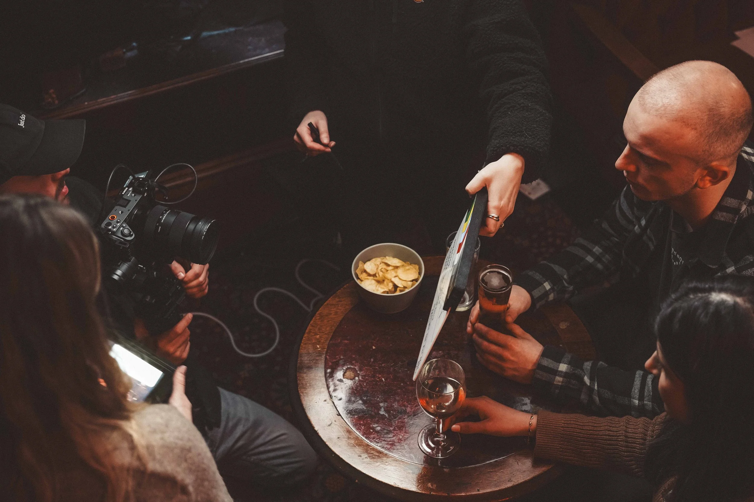 People sitting around a table with snacks and drinks, a person holding a camera, and one person wearing a black cap and a black sweater.