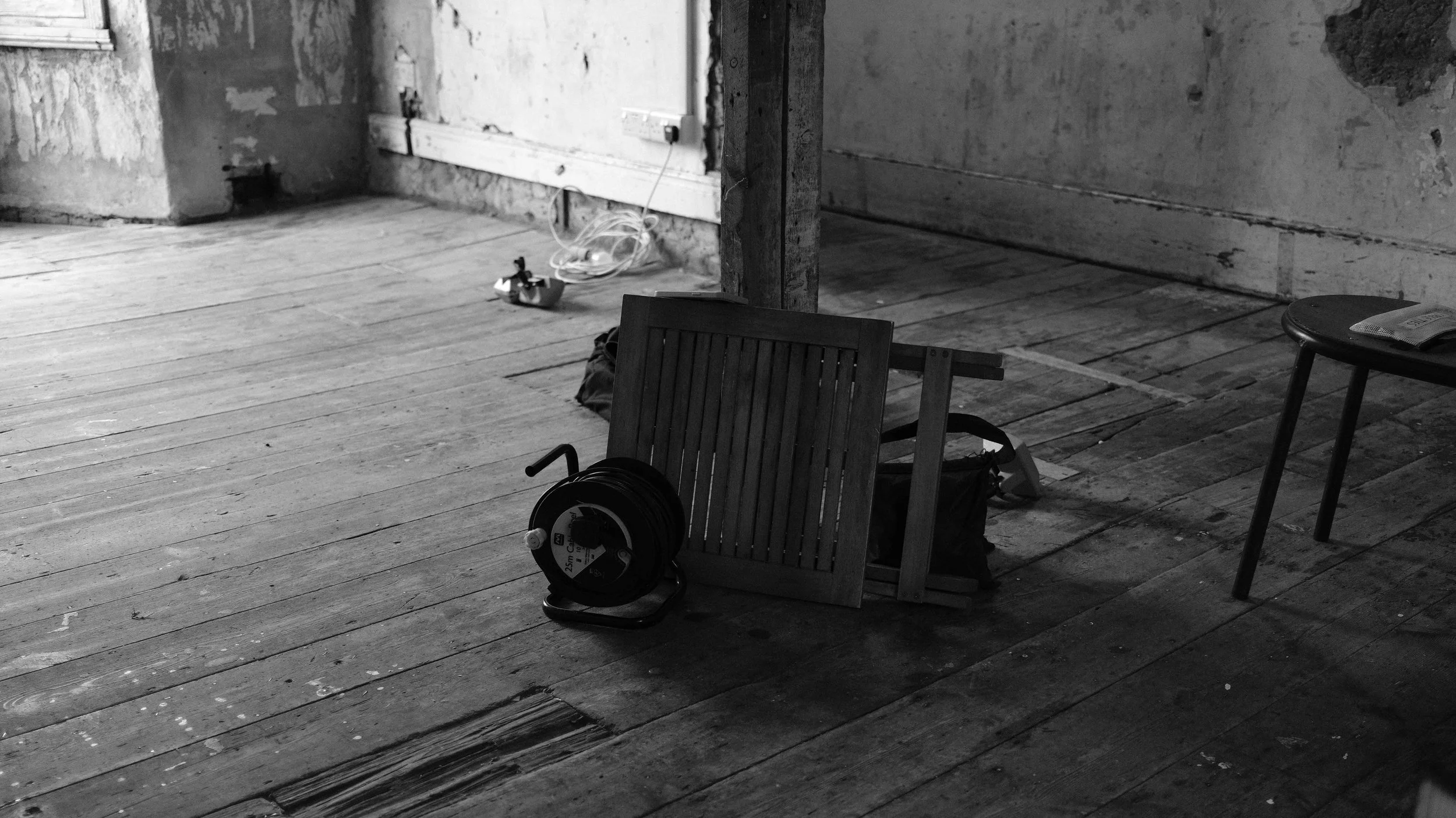 A black and white photo of a rustic, unfinished room with wooden floorboards. There is a wooden chair, a small round stool with a laptop, and tools such as a coiled extension cord and a small handheld device on the floor. The walls appear worn, and t