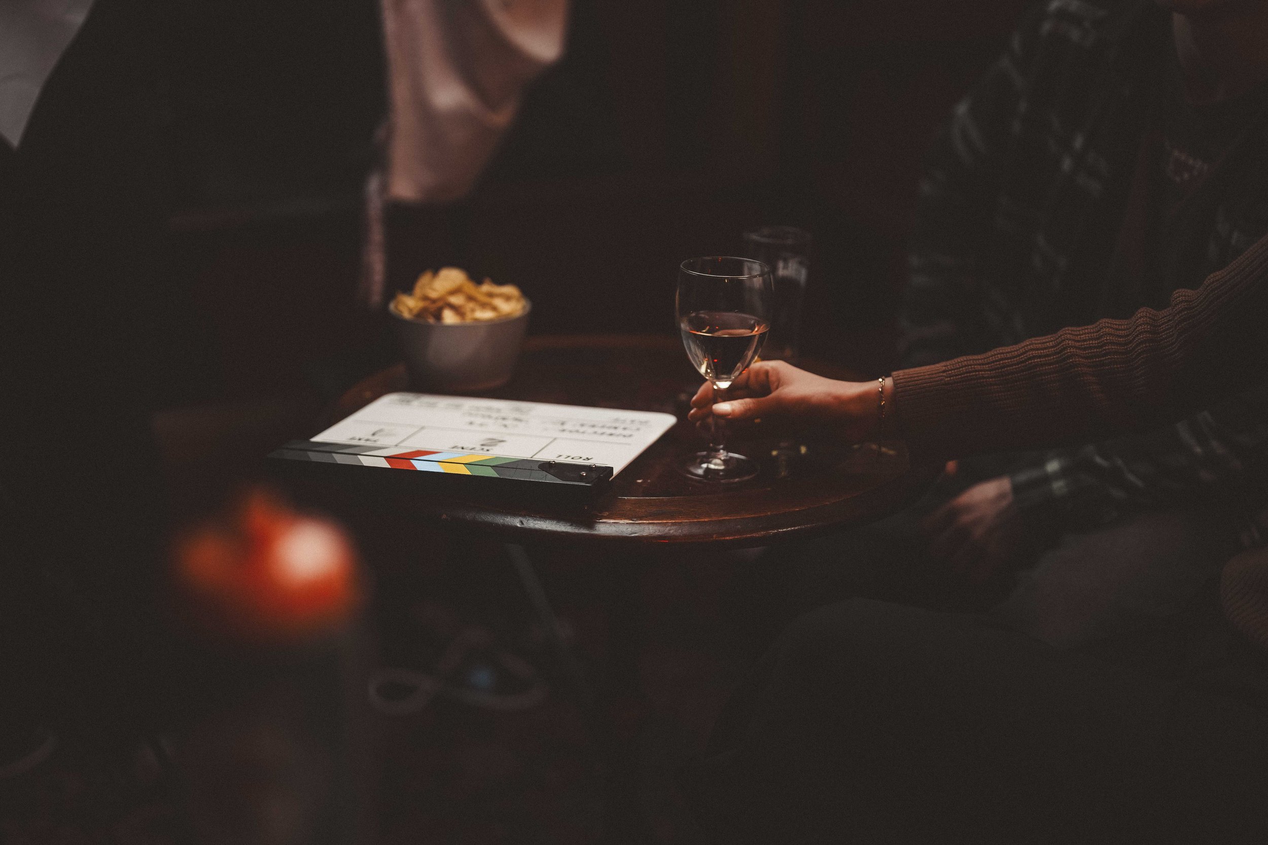 A person holding a glass of pink wine at a dark table with a small bowl of food and a clapperboard.
