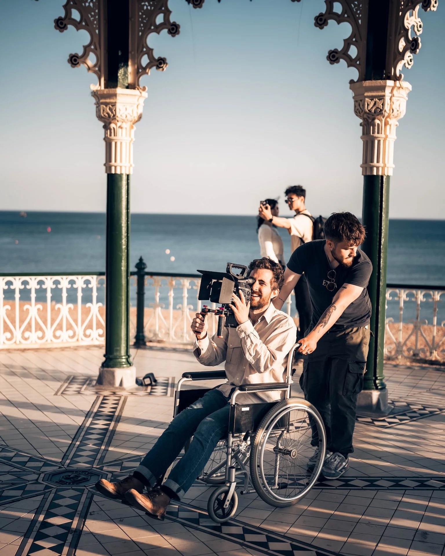 Three men on a decorative seaside pavilion with ornate columns, one sitting in a wheelchair holding a camera, and two others standing nearby, one assisting him. In the background, the sea and sky are visible.