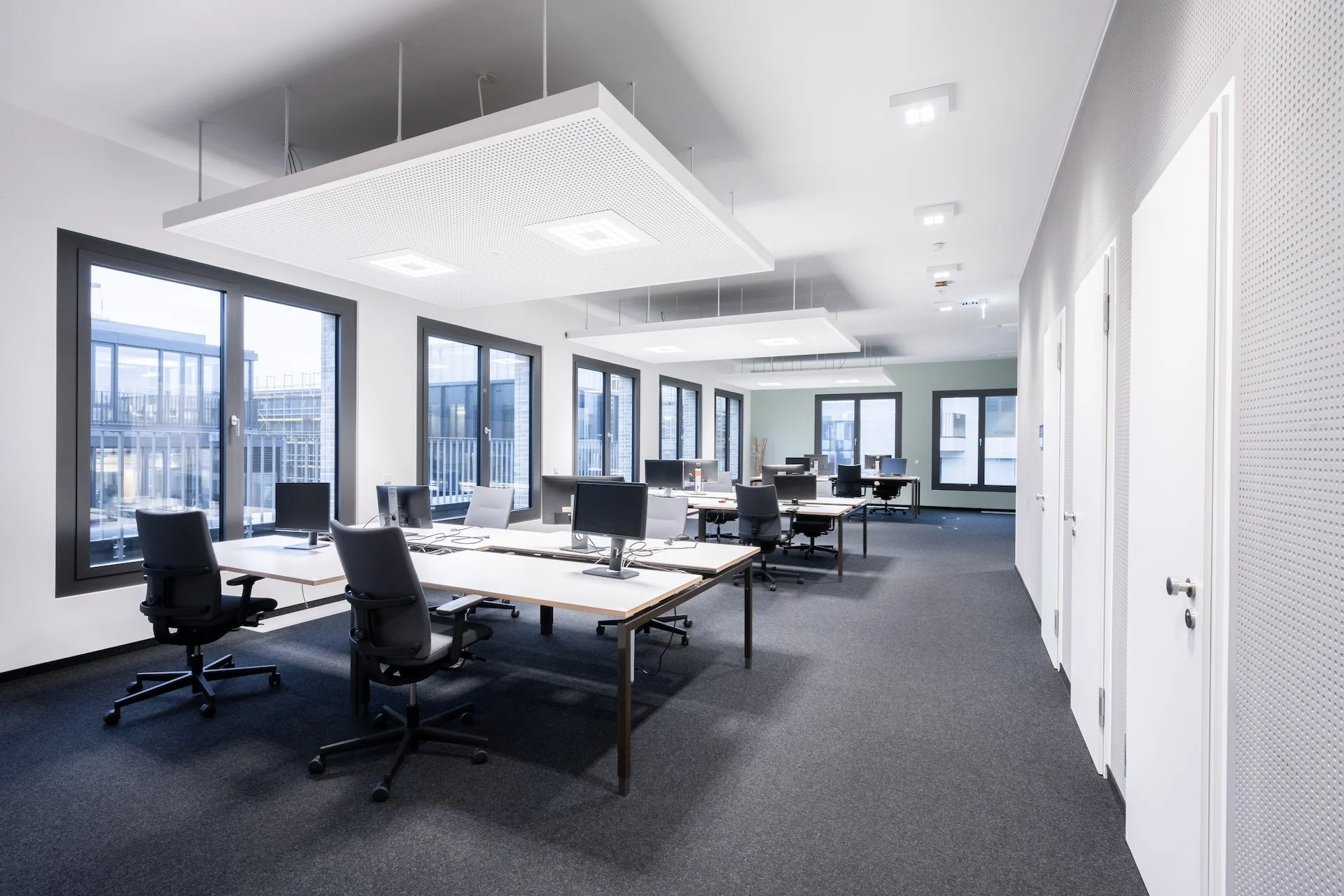 Empty modern office with desks, chairs, desktop computers, large windows, and ceiling lights.