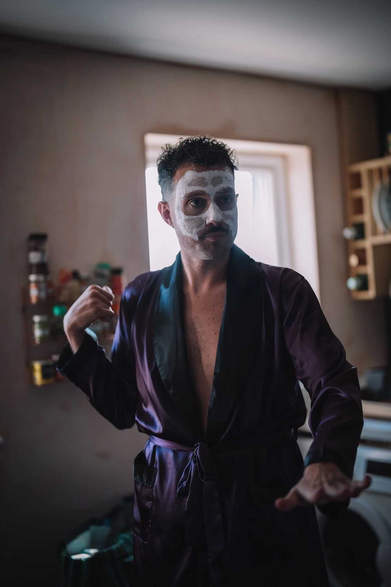 A man with a white face mask makes it look like a skull, wearing a dark purple satin robe, standing in a room with a window behind him, kitchen shelves with jars on the side.