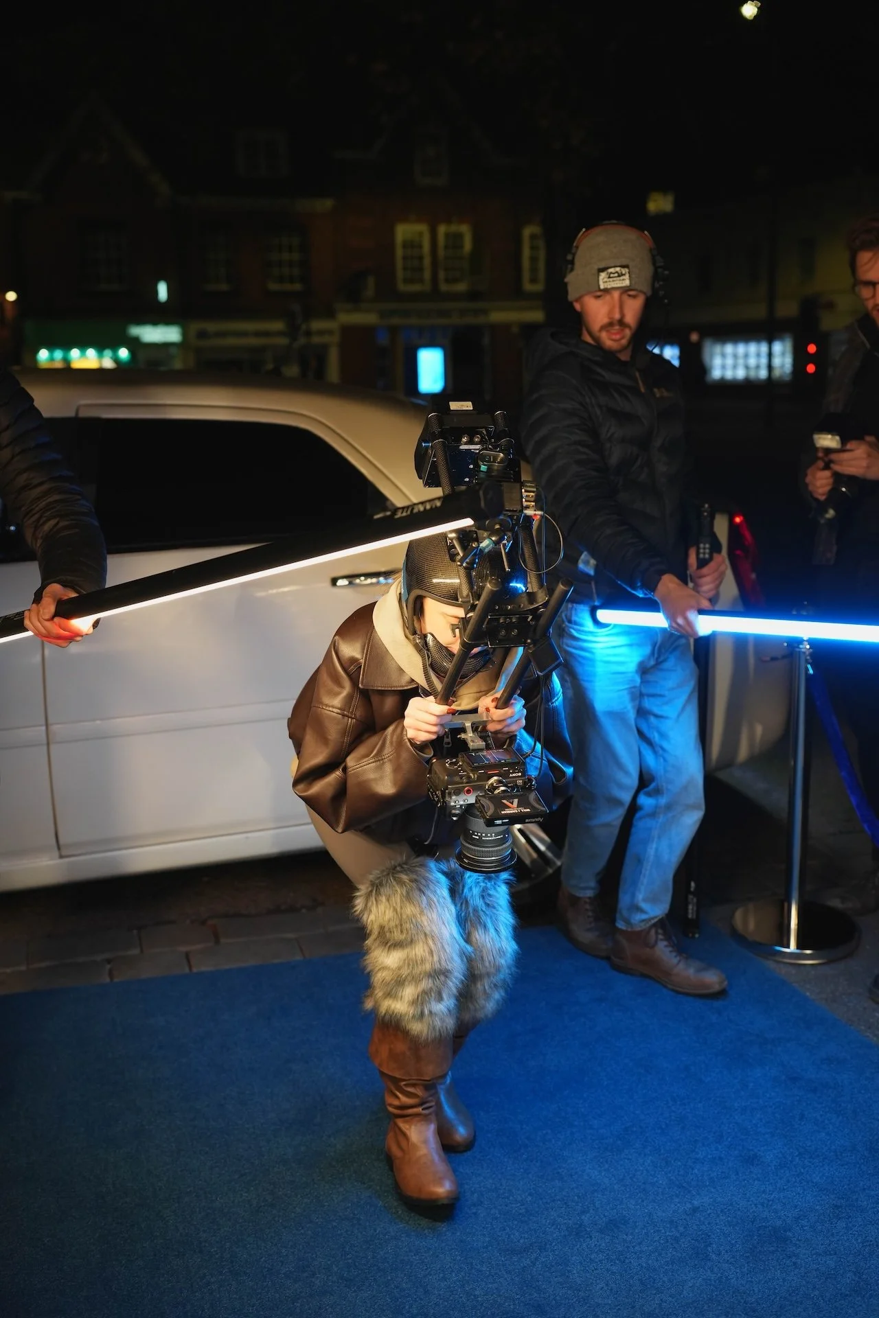 Filmmakers and crew setting up filming equipment on a city street at night, with a person in a leather jacket, furry boots, and hat operating a camera, and others holding lights and equipment.