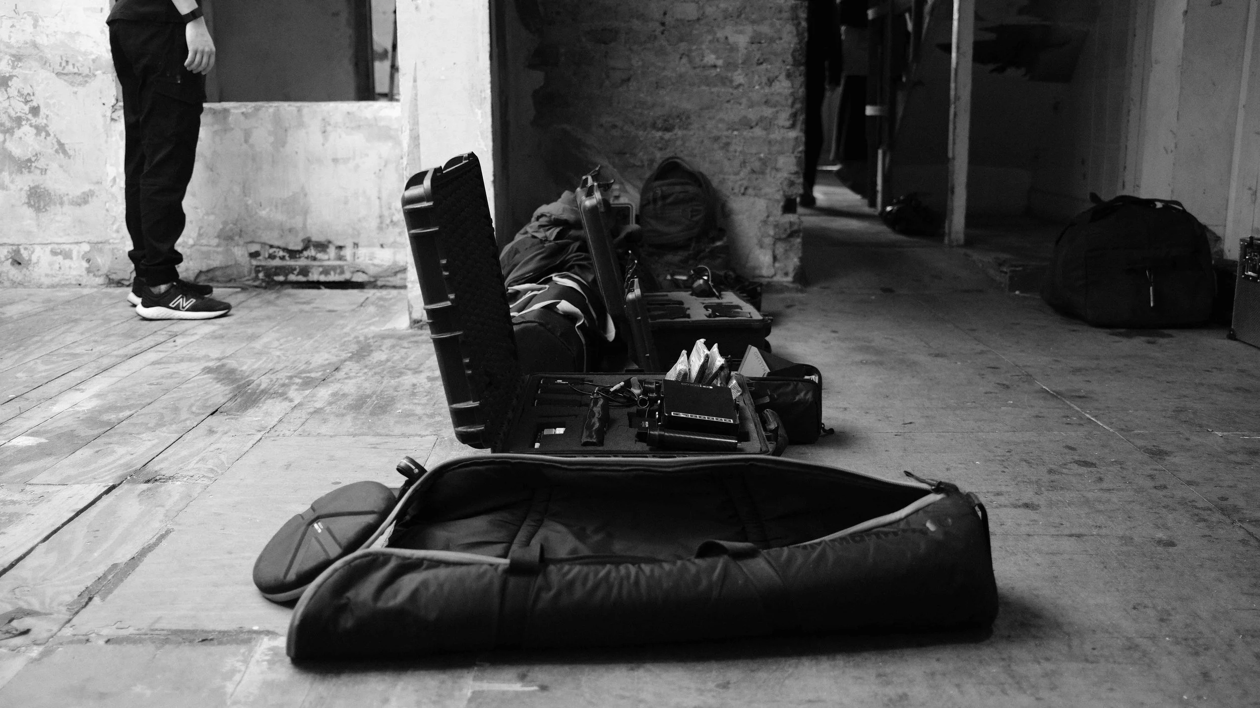 Open black guitar case on a wooden floor with items inside, surrounded by other bags and equipment in a room with brick and concrete walls.