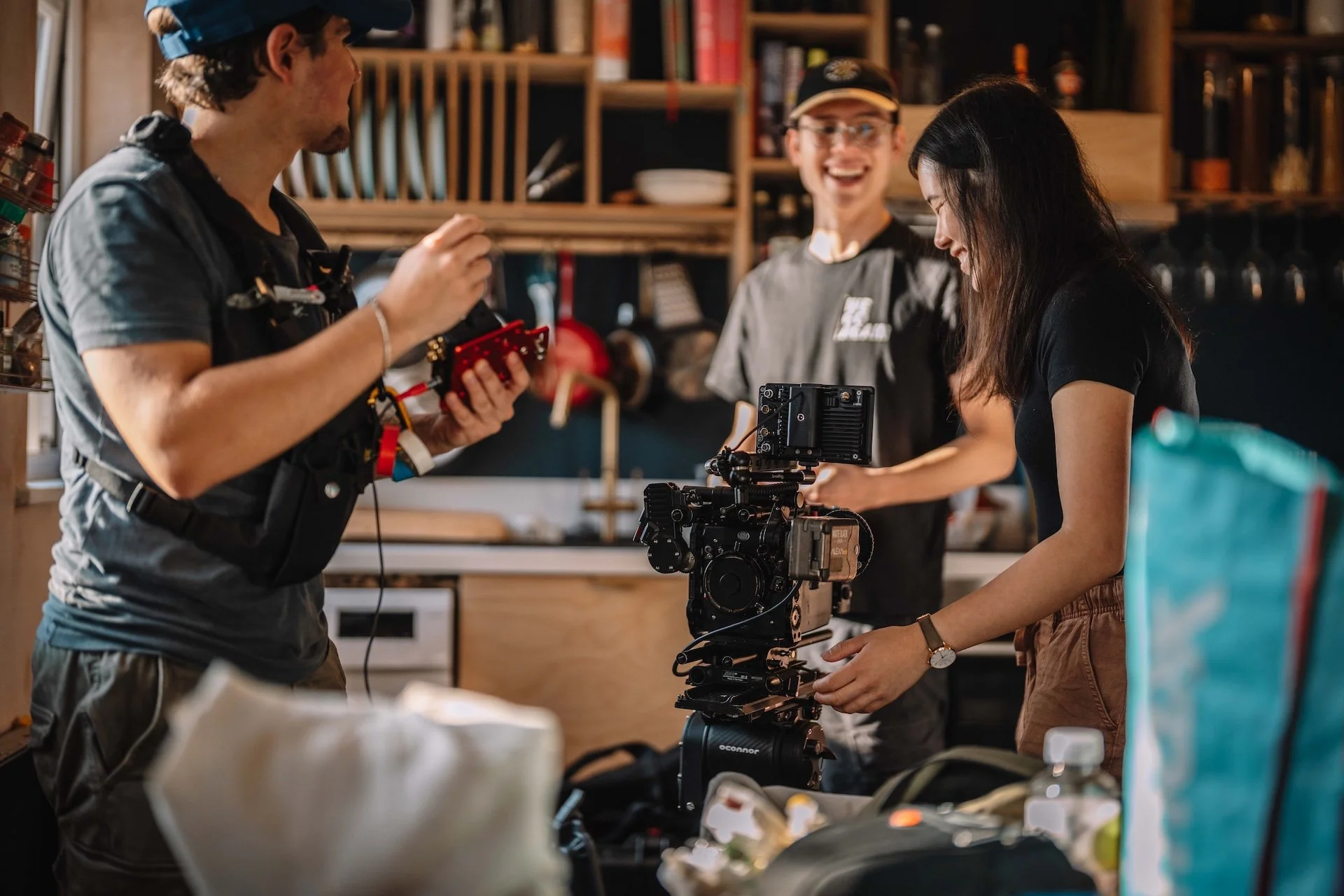 Three people working together on a film or video shoot in a cozy, organized kitchen or workshop. One person is adjusting equipment, another is operating a camera rig, and the third is smiling in the background.