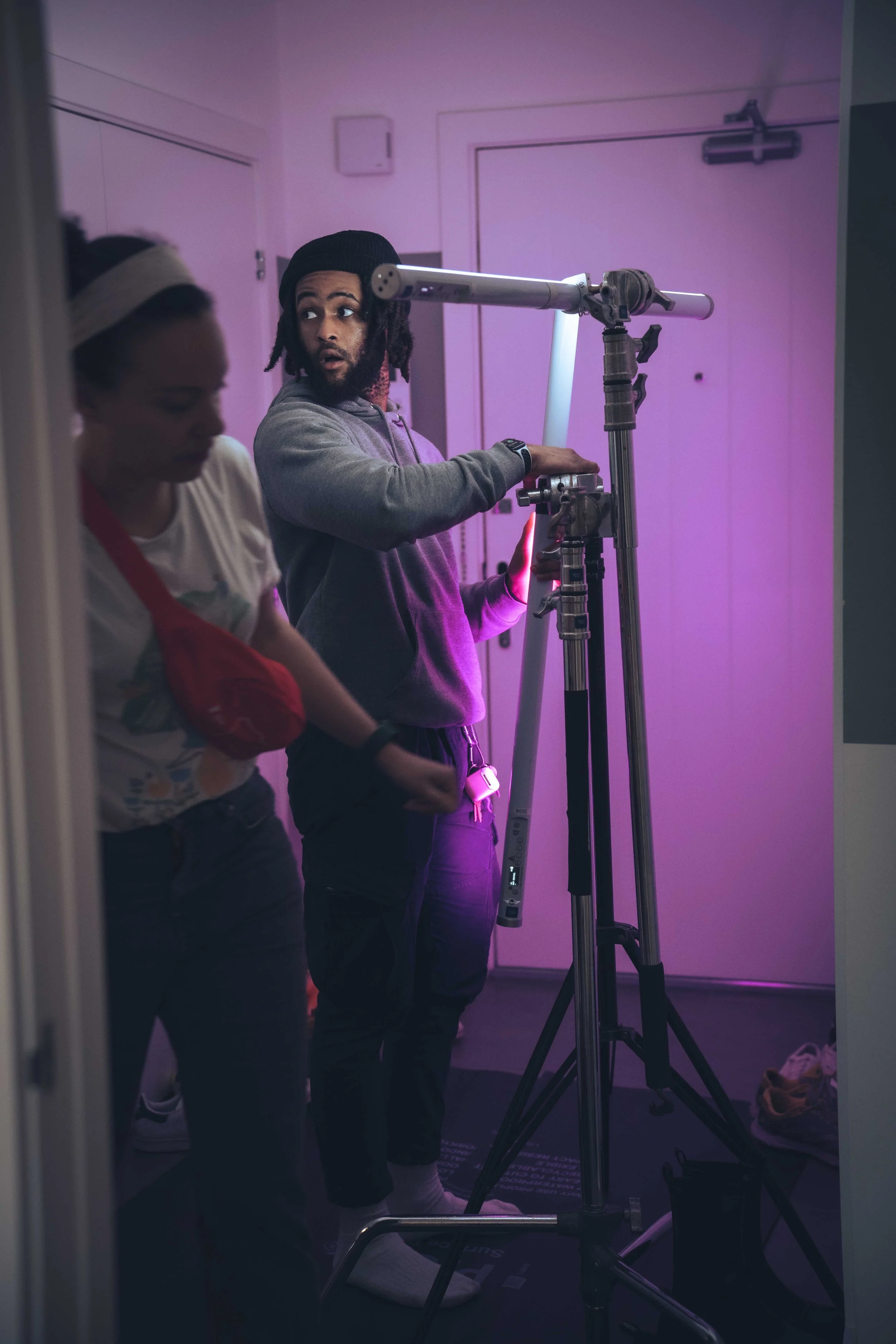 Two people in a dimly lit room with purple lighting; one person is adjusting a telescope mounted on a tripod, while the other person, a woman in a white shirt and red apron, appears to be looking down or away.